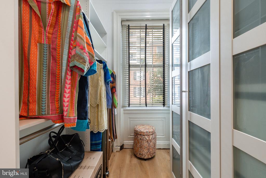 Walk-in closet with colorful hanging clothes, a patterned ottoman, and a window with blinds.
