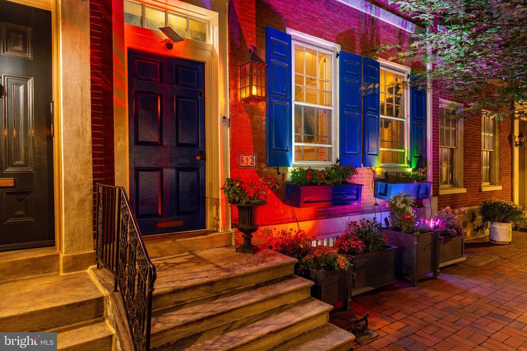 Brick rowhouse exterior at night with blue shutters, illuminated windows, and planted window boxes.