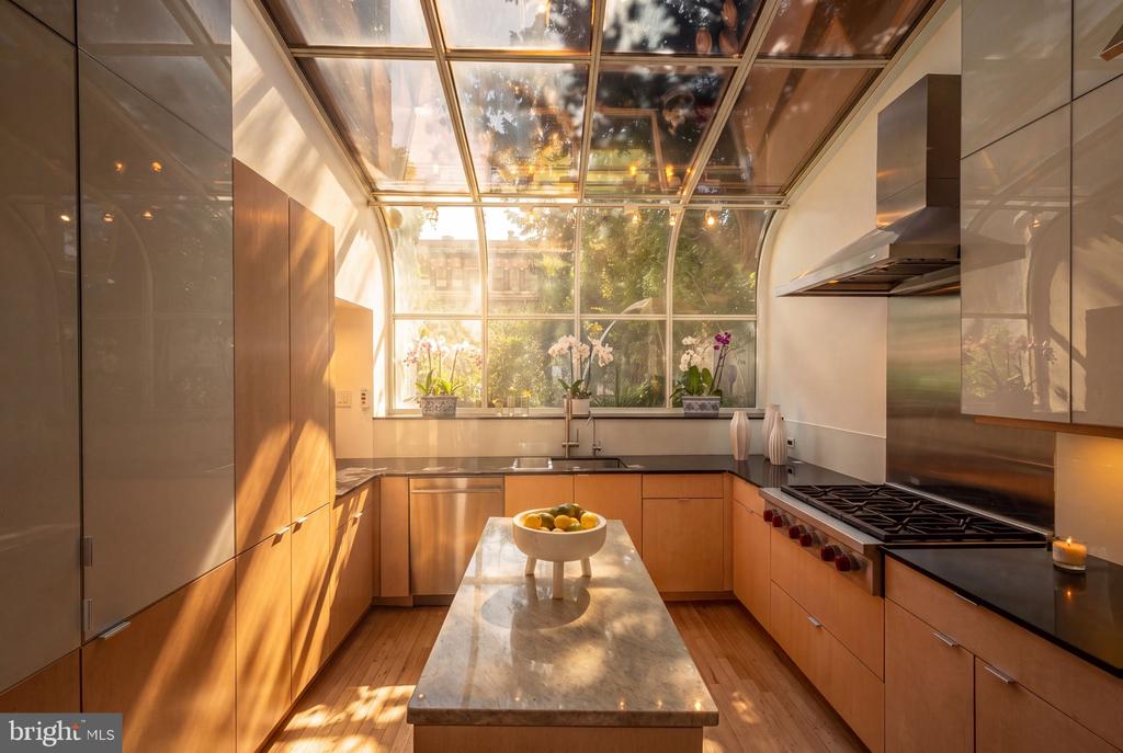 Modern kitchen with wood cabinetry, marble island, and glass ceiling overlooking greenery.