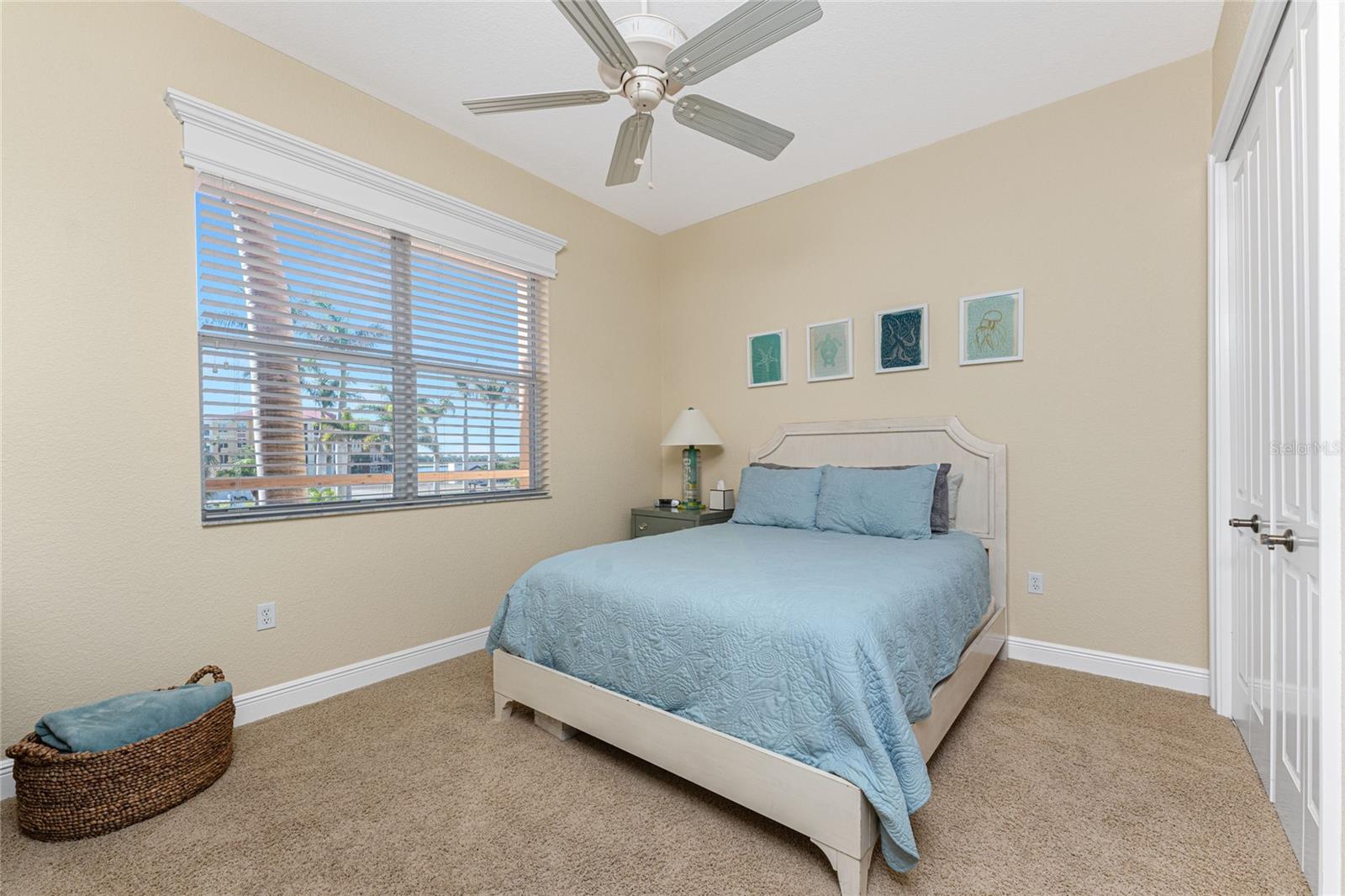 Bedroom with pale blue bedding, sea-life artwork, and a wide window overlooking palm trees and water.