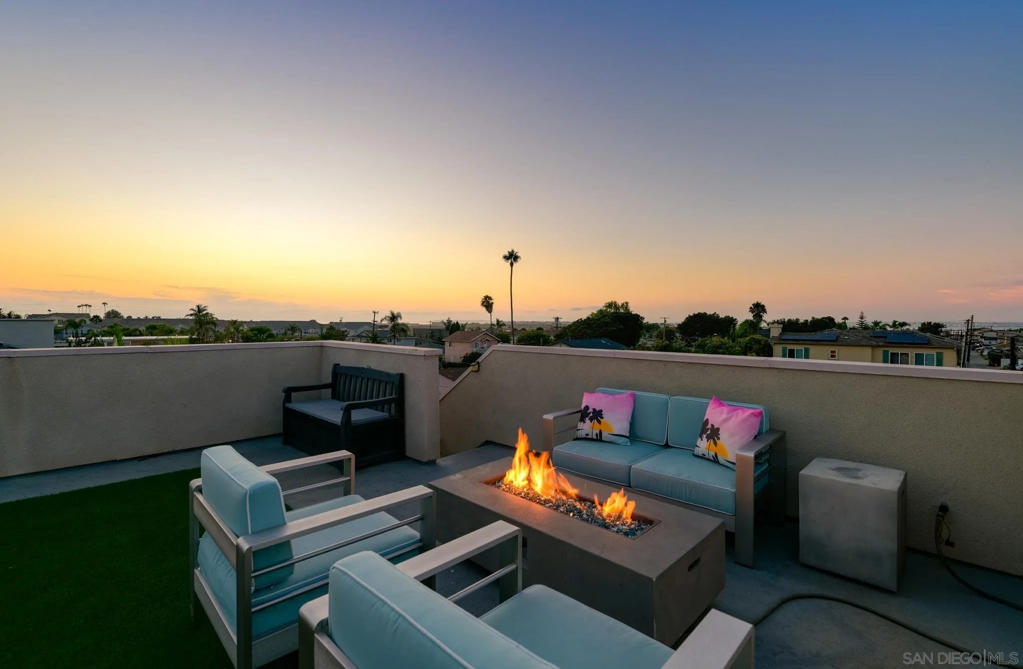 Rooftop patio seating around a gas fire pit at sunset, with palm trees and neighborhood views.