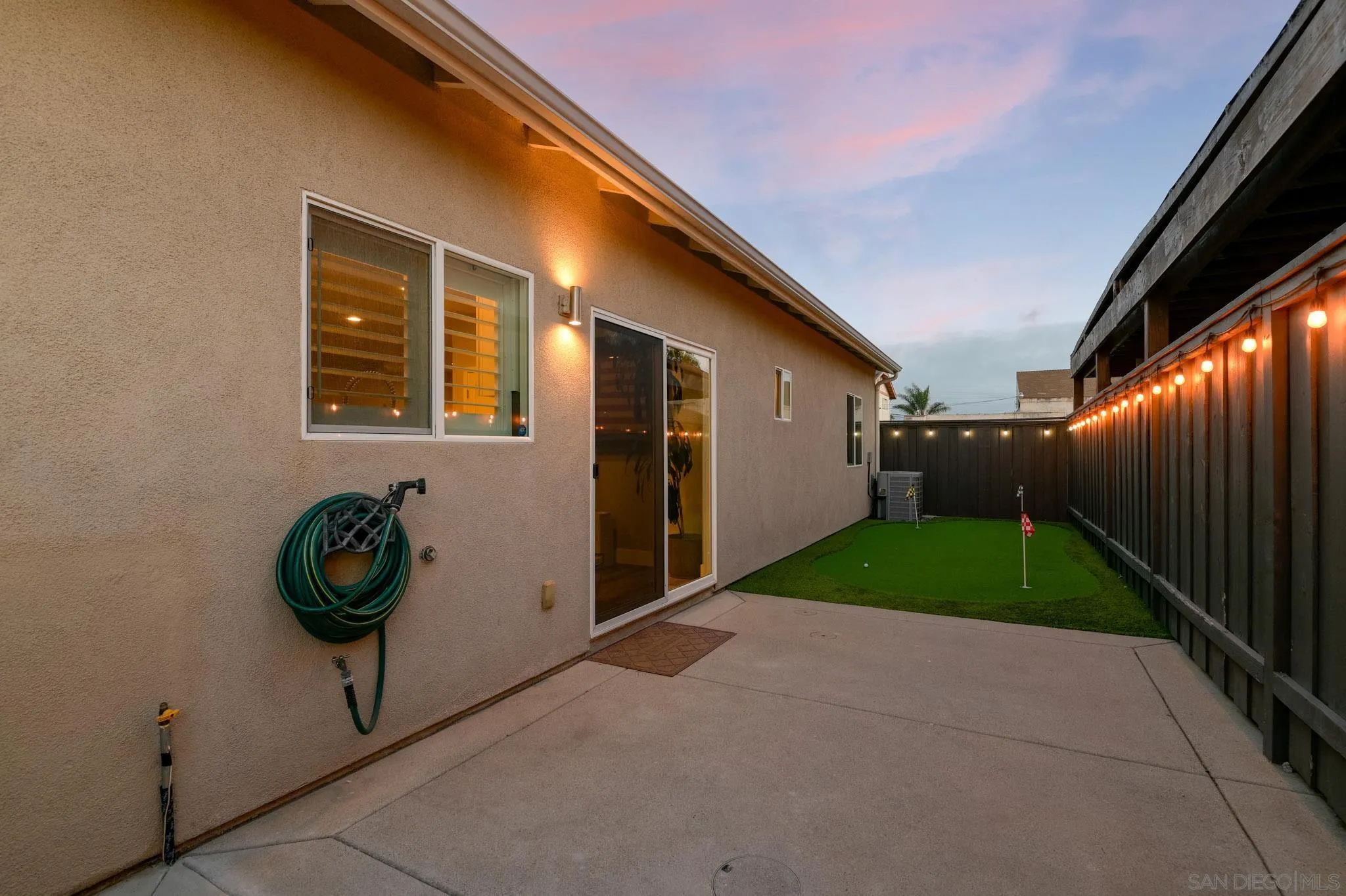 Side patio with a fenced artificial-turf putting green and glowing string lights at dusk.