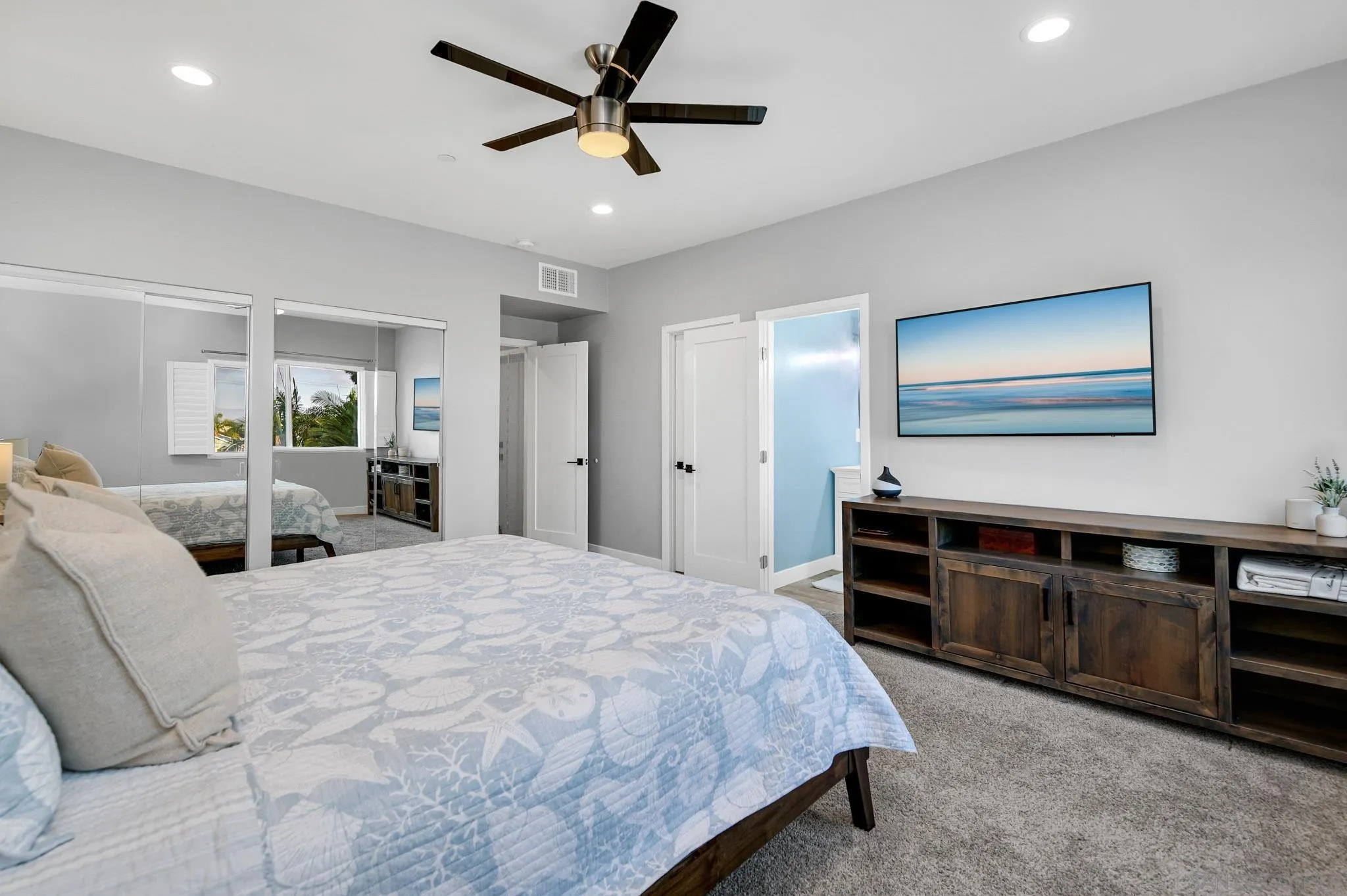 Staged bedroom with mirrored closet doors and a dark wood media console beneath a wall-mounted TV.