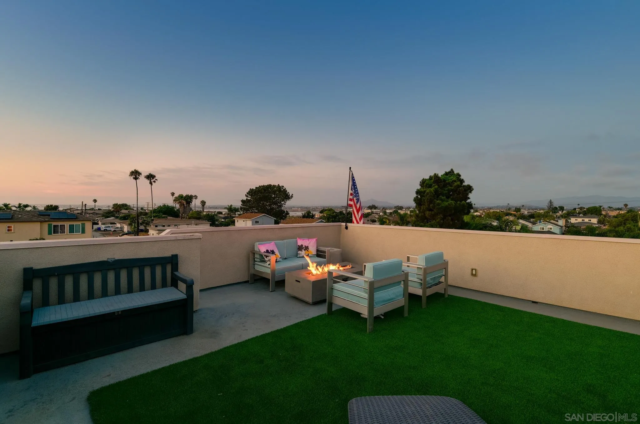 Rooftop terrace with a fire table, lounge seating, artificial grass, and neighborhood views at dusk.