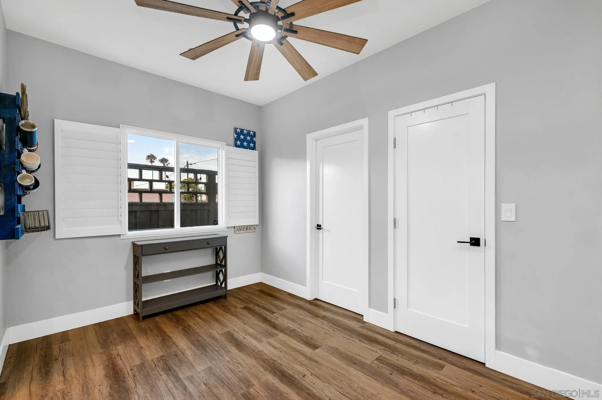 Bright room with wood-look floors, a ceiling fan, shuttered window, and two white doors.