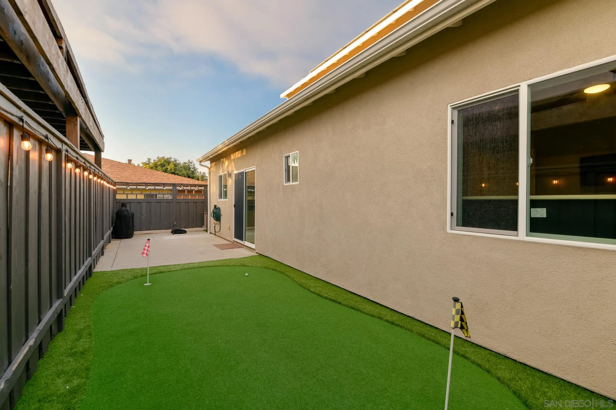 Backyard putting green beside a beige stucco house, with golf flags, a concrete patio, and string lights.