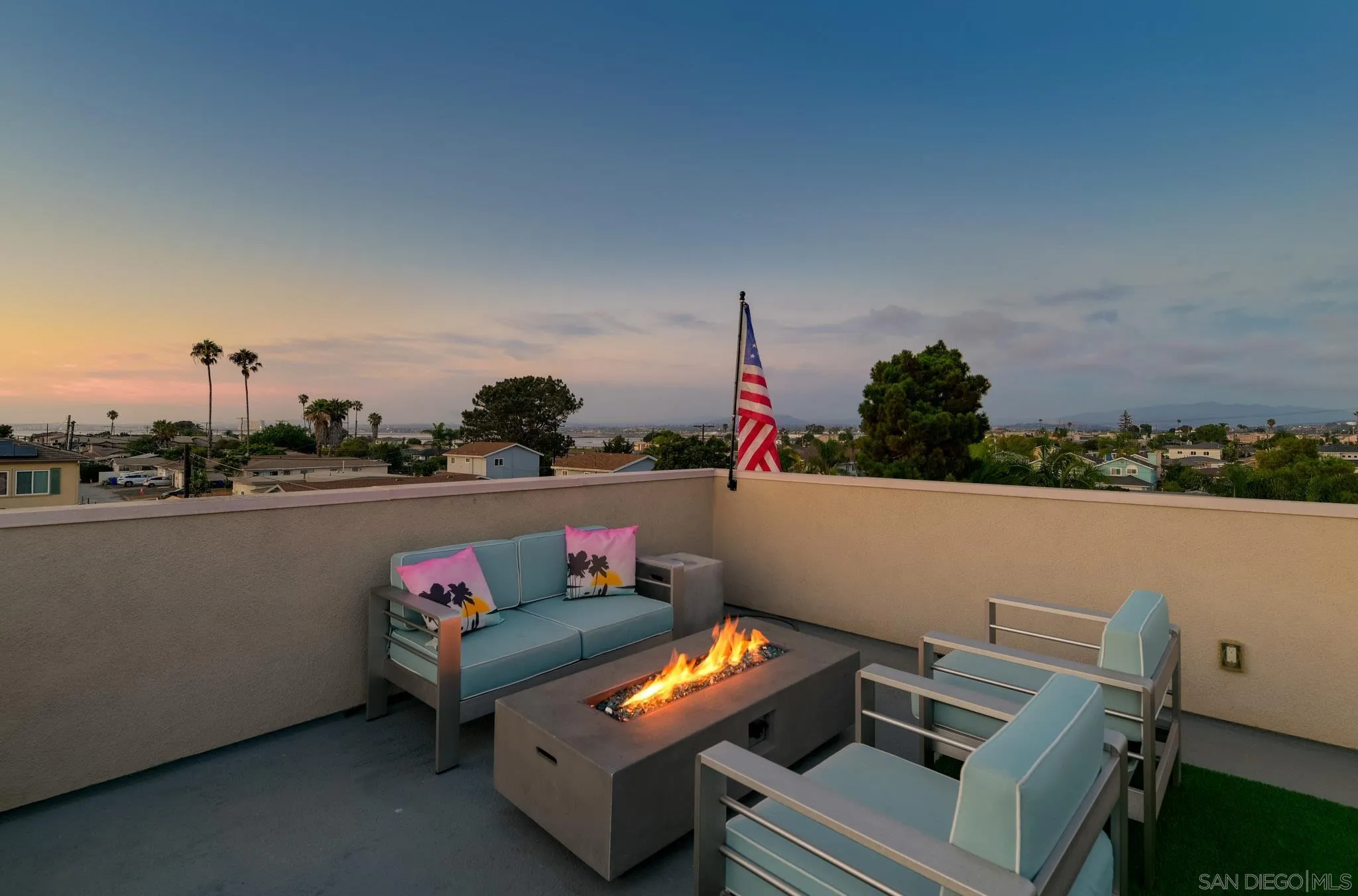 Rooftop terrace with blue seating, a lit fire pit, and neighborhood views at sunset.