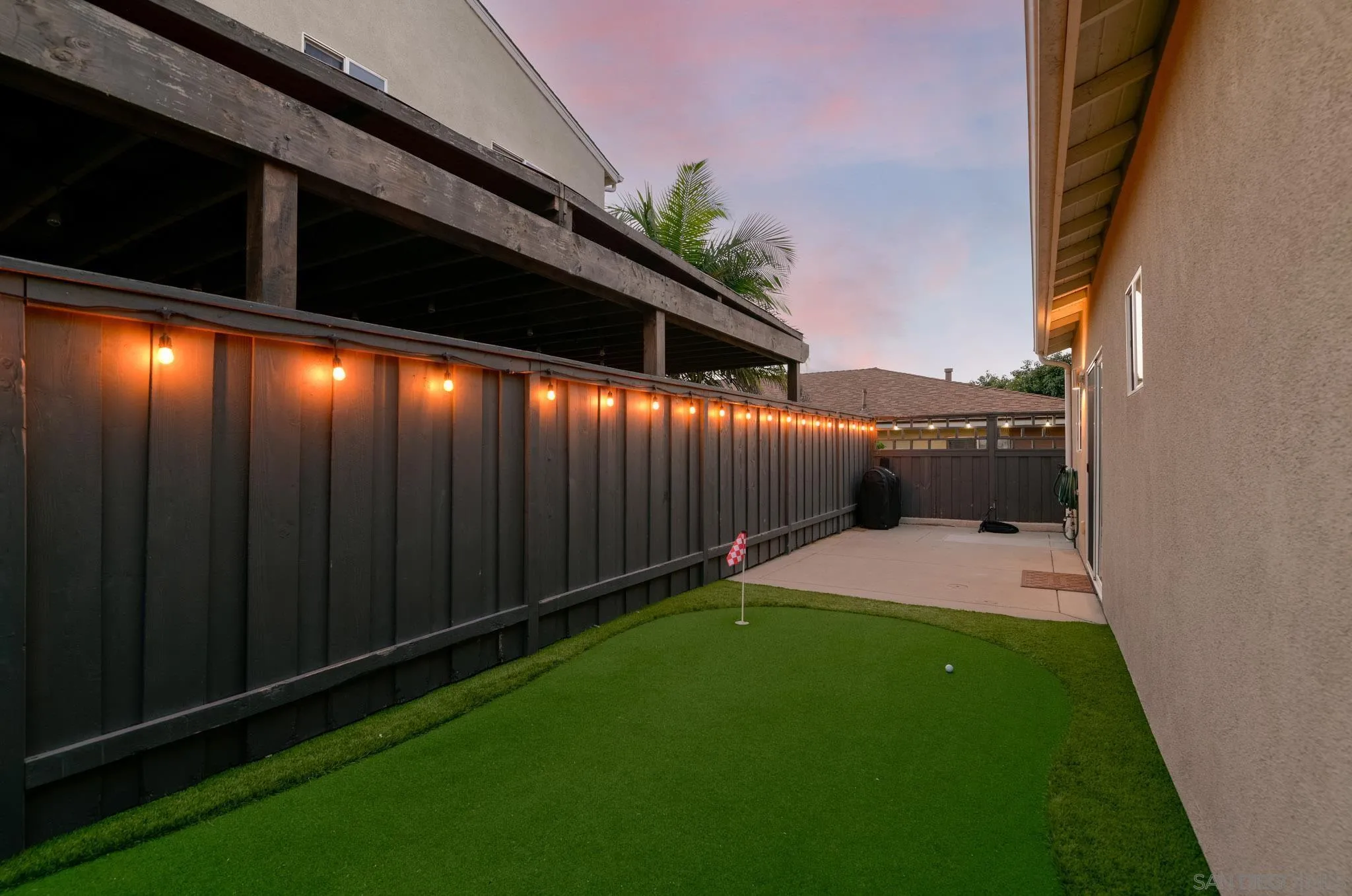 Backyard putting green beside a dark fence strung with warm lights and a raised deck.