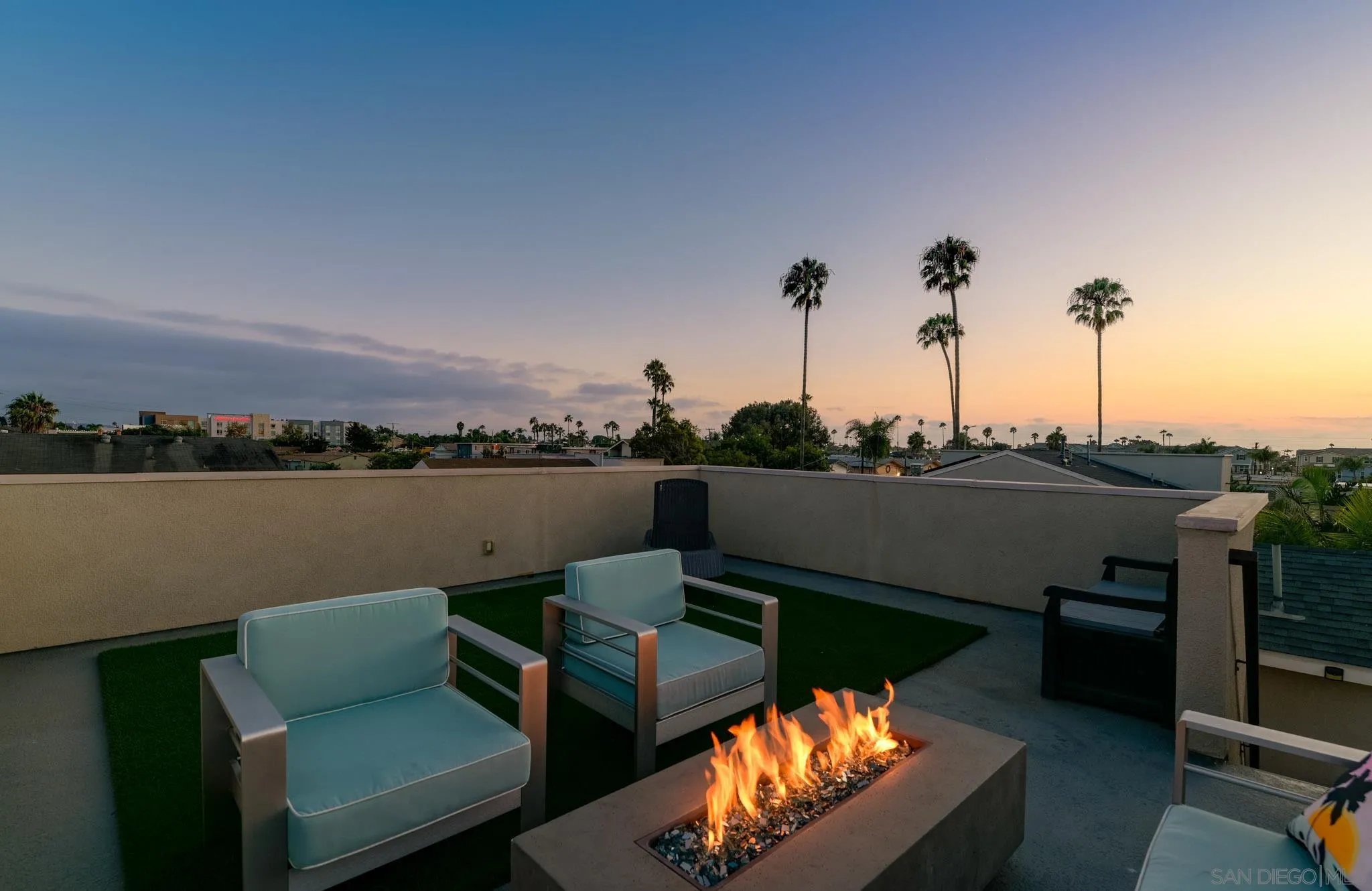 Rooftop terrace with lounge chairs and a gas fire pit overlooking palm trees at sunset.