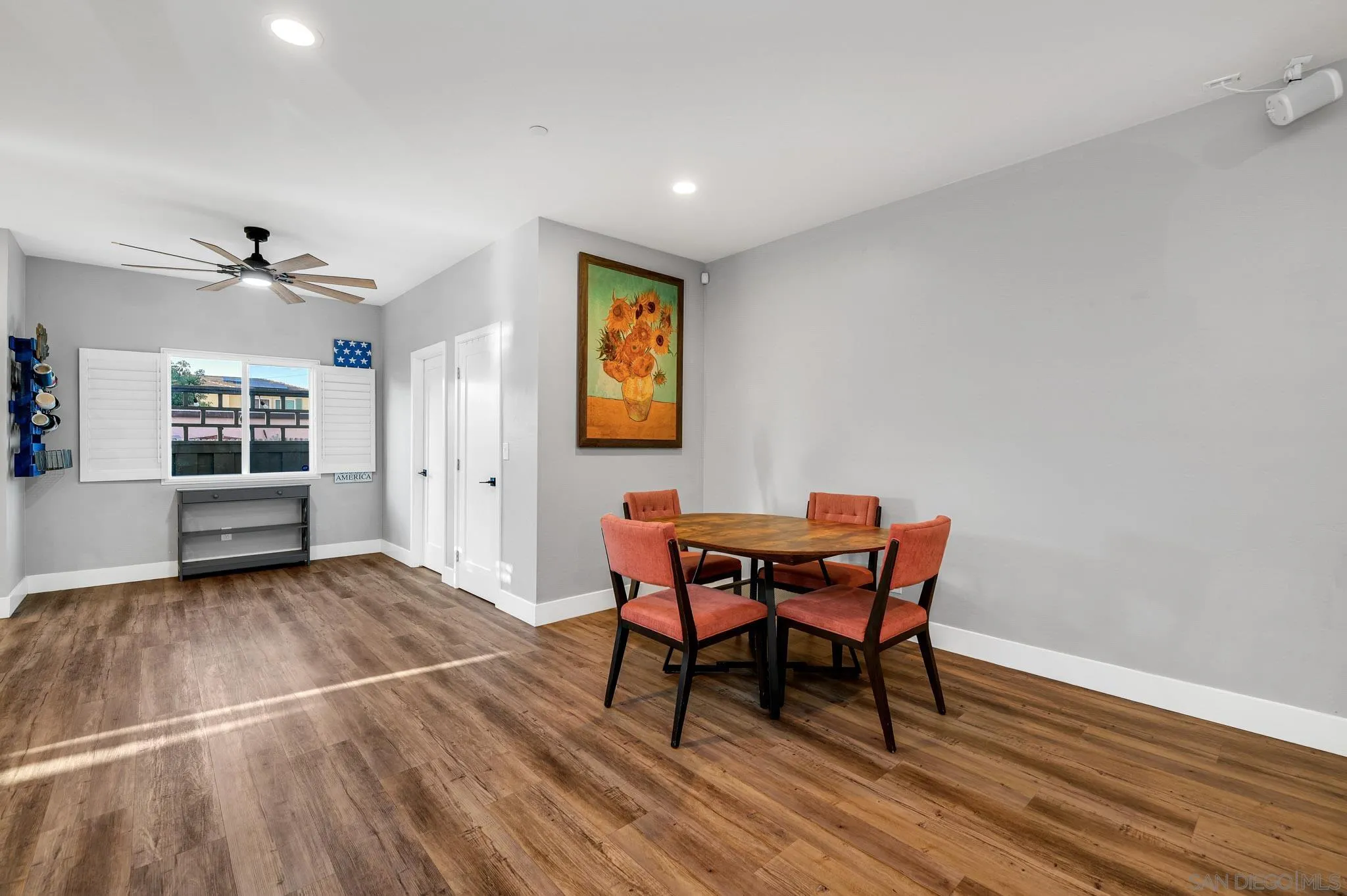 Dining area with a round wood table, coral chairs, shuttered window, and sunflower painting.
