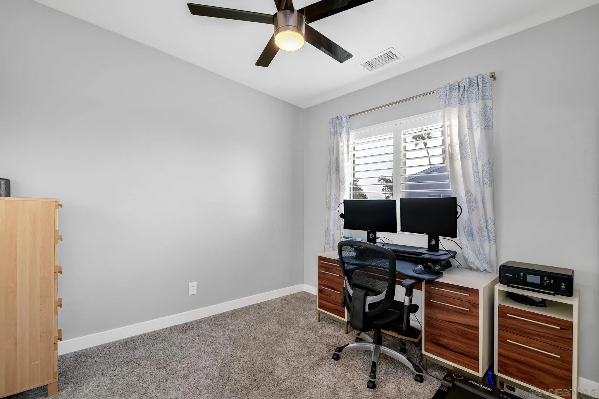 Home office with a dual-monitor desk beneath a shuttered window and a ceiling fan.