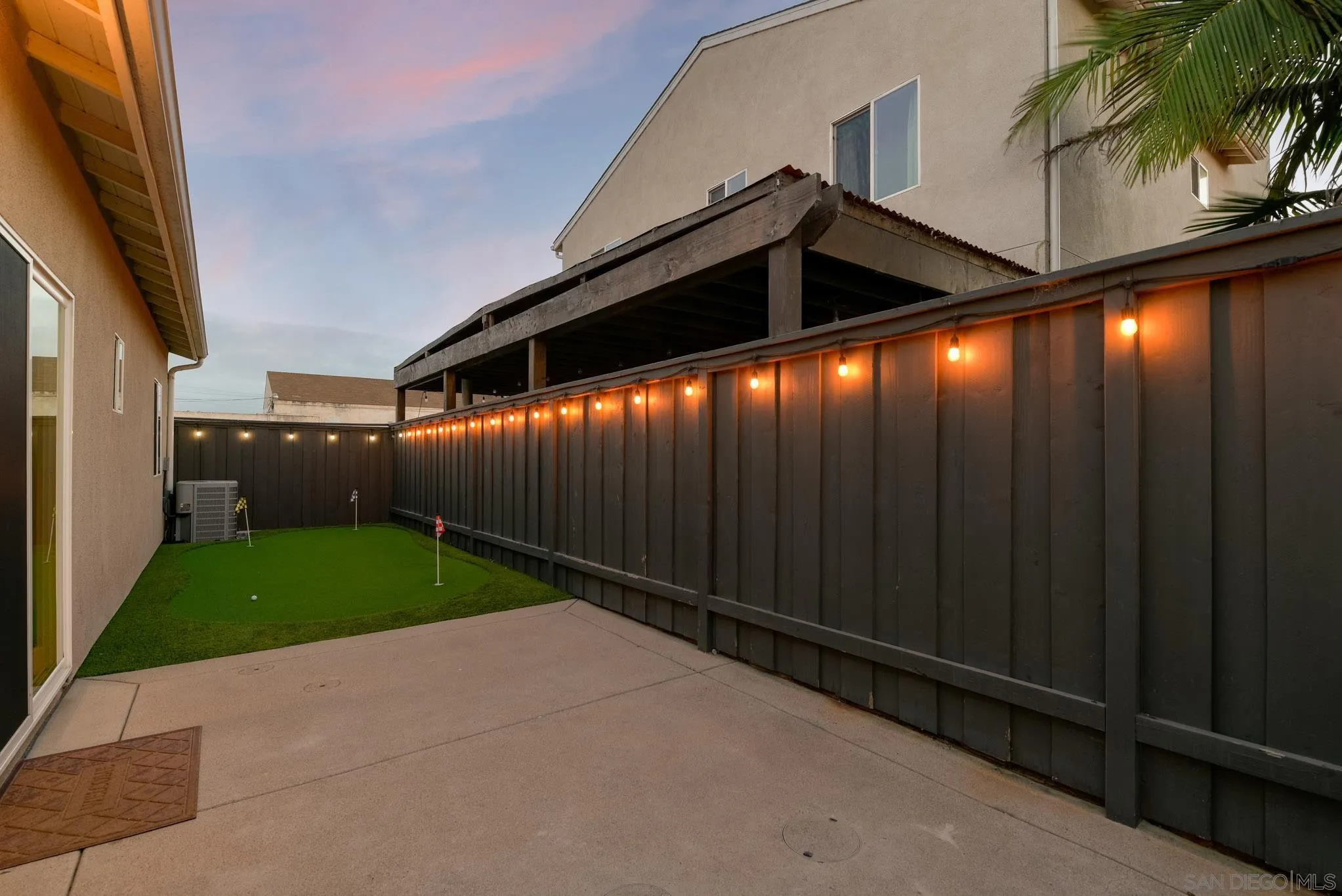 Backyard putting green beside a concrete patio, with string lights along the privacy fence at dusk.
