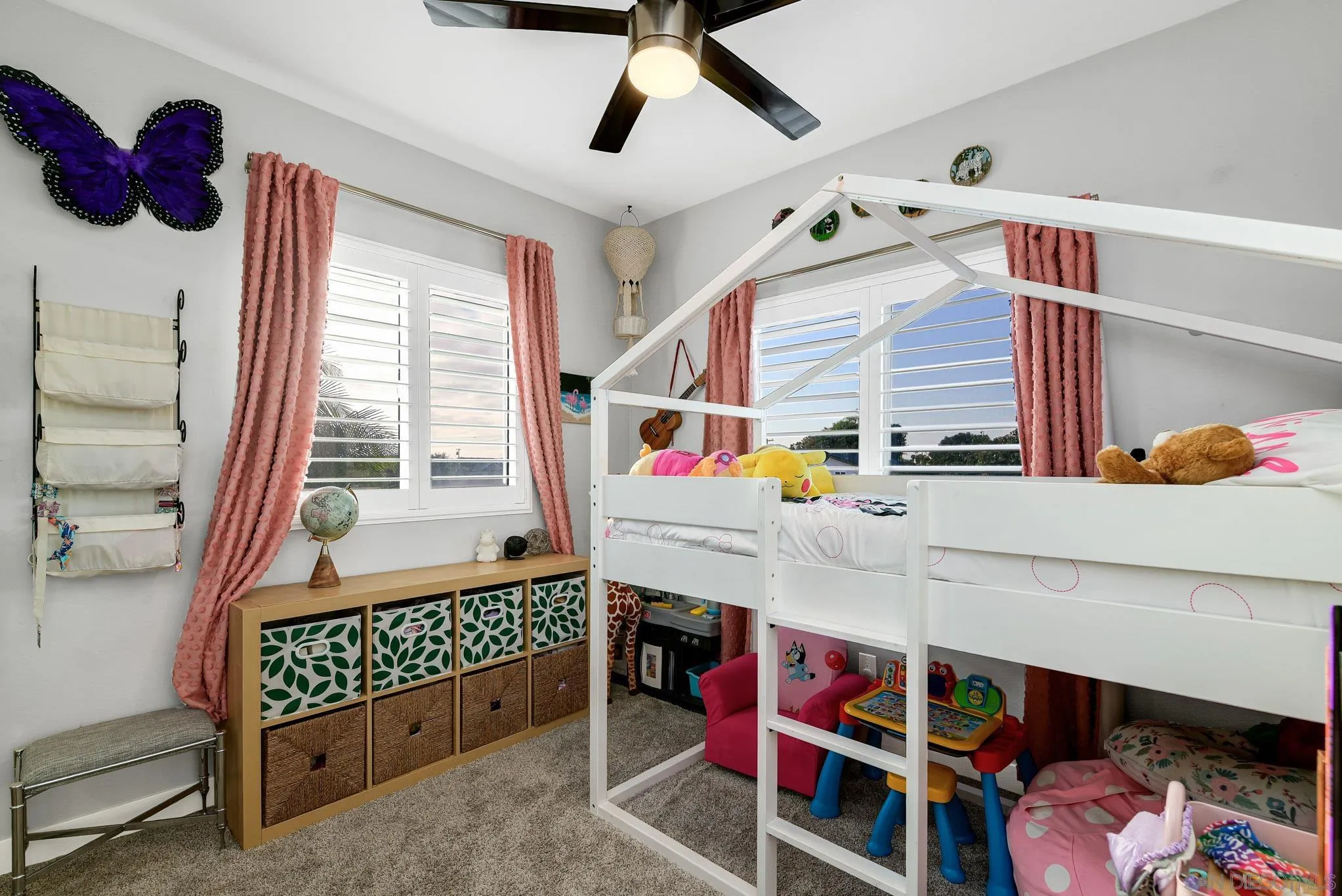 Children’s bedroom with a white house-frame bunk bed, toys, pink curtains, and shuttered windows.