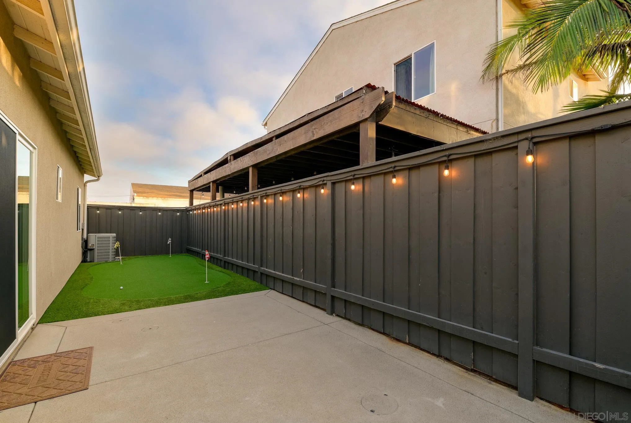 Fenced backyard patio with an artificial-turf putting green, golf flags, and warm string lights.
