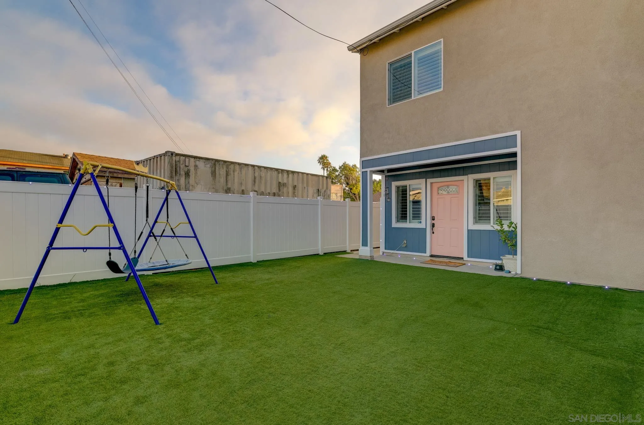 Backyard with a wide artificial-turf lawn, white privacy fence, and children's swing set beside the home.