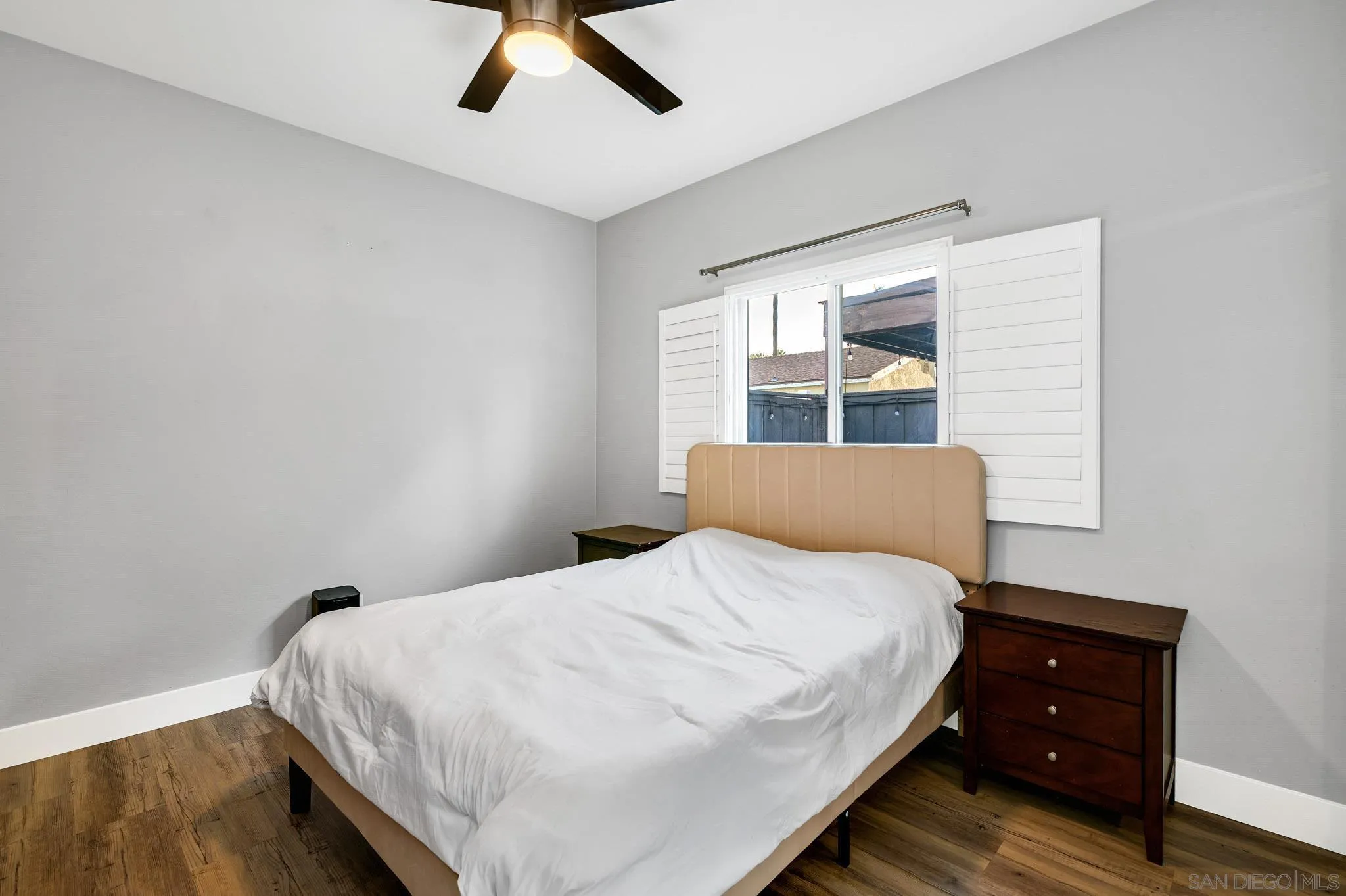 Bedroom with a white-covered bed, tan upholstered headboard, shuttered window, and wood-look flooring.