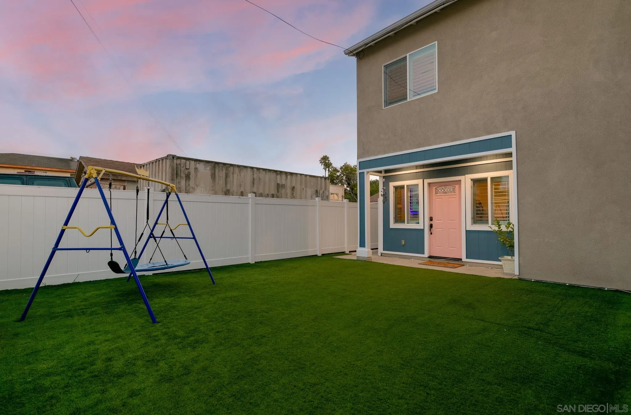 Home’s backyard with a wide lawn, swing set, white fence, and pink entry door.