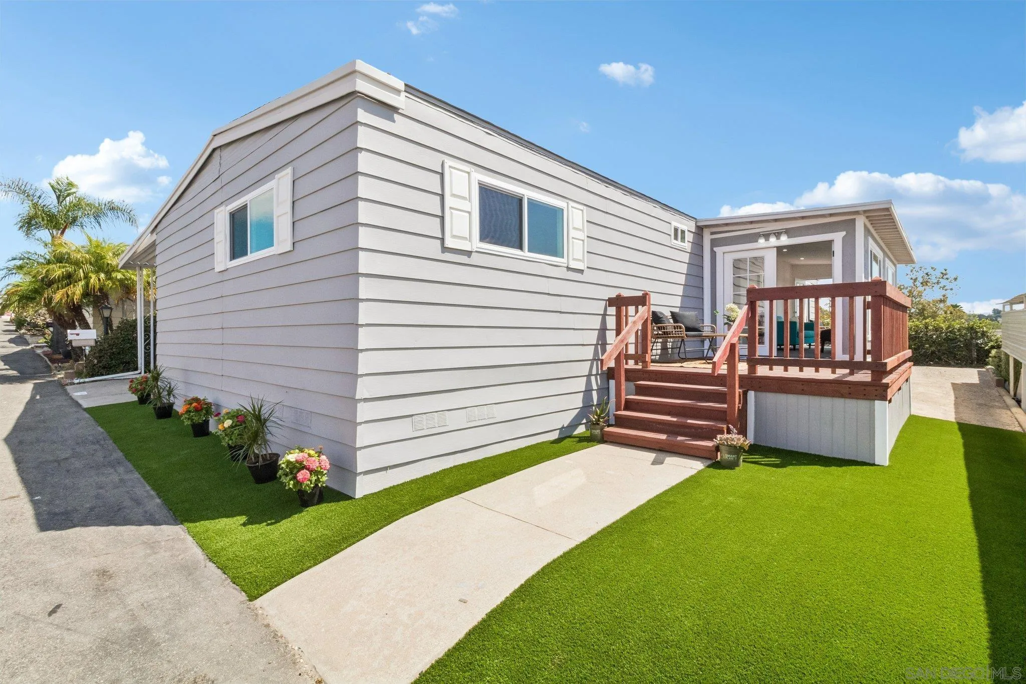 Gray-sided home with a raised reddish-brown deck, patio seating, potted flowers, and green lawn.