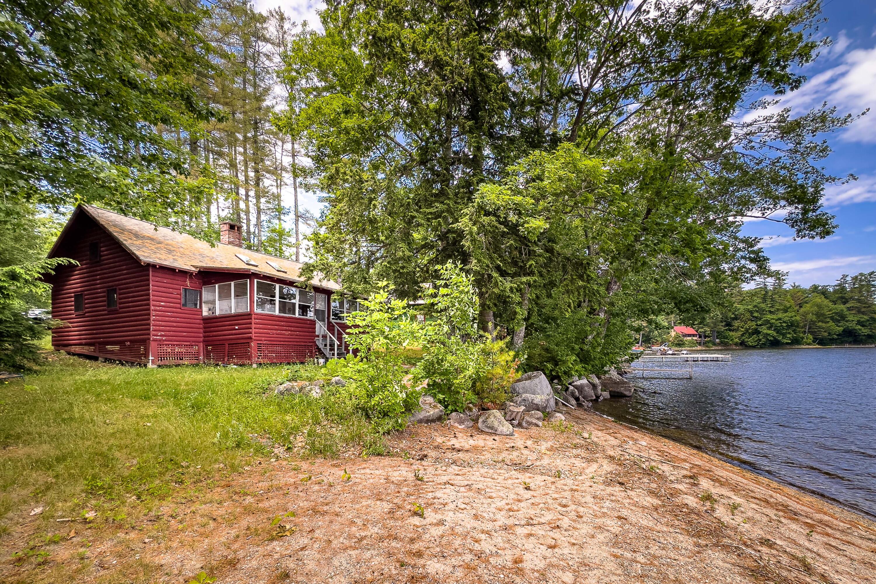 Red log cottage on a wooded lakeshore beside a rocky beach and small dock.