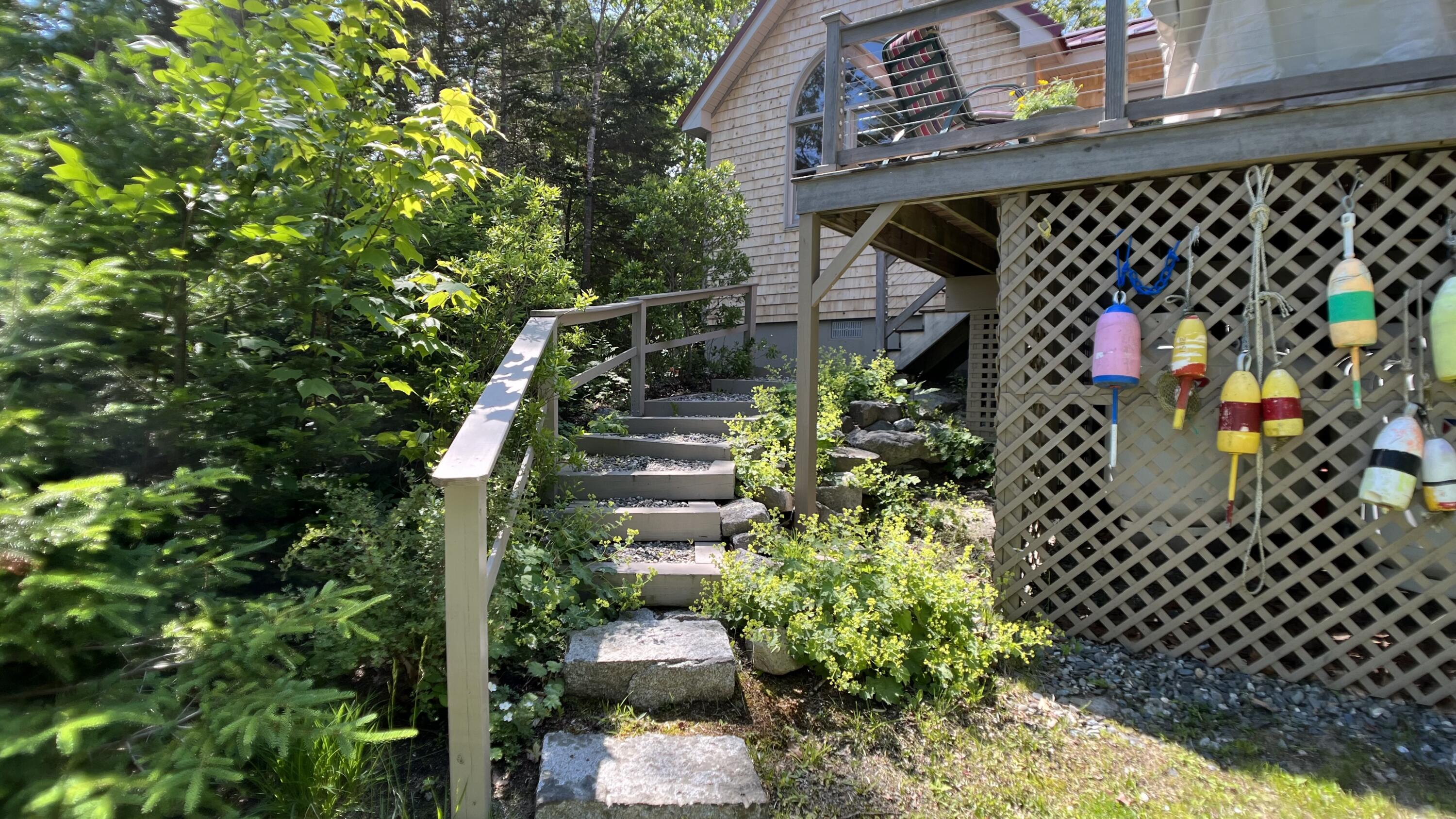 Shingled house with a raised deck, stone steps, hanging fishing buoys, and lush garden landscaping.