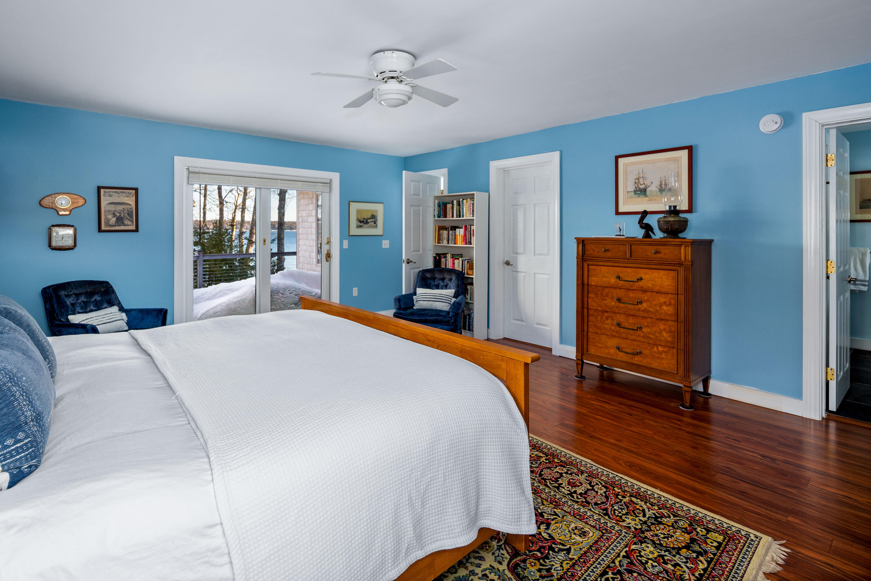 Blue bedroom with a white-covered bed, wooden dresser, and sliding doors overlooking water and snow.