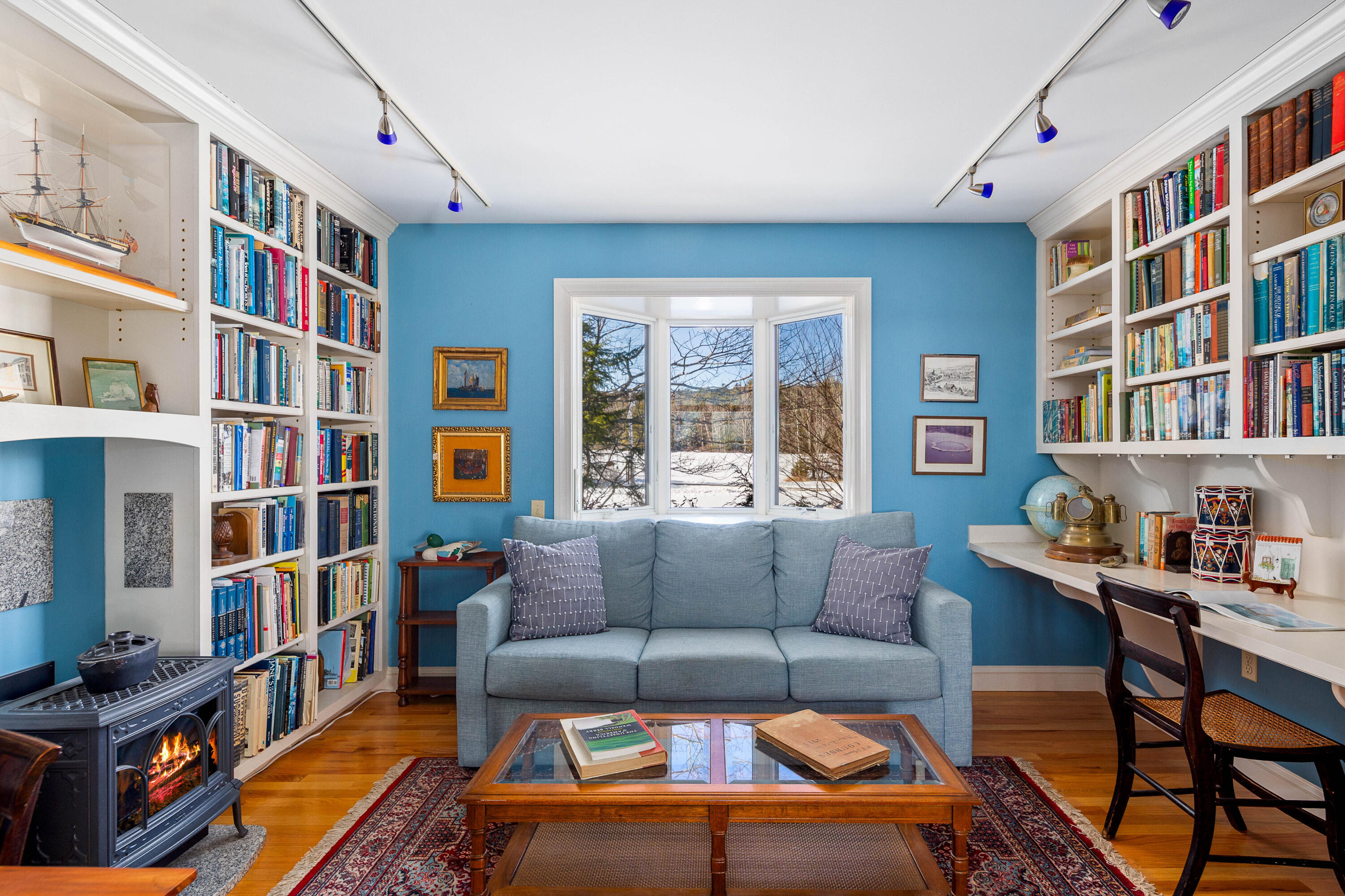Furnished blue library with wall-to-wall bookshelves, a bay window, gray sofa, and lit wood stove.