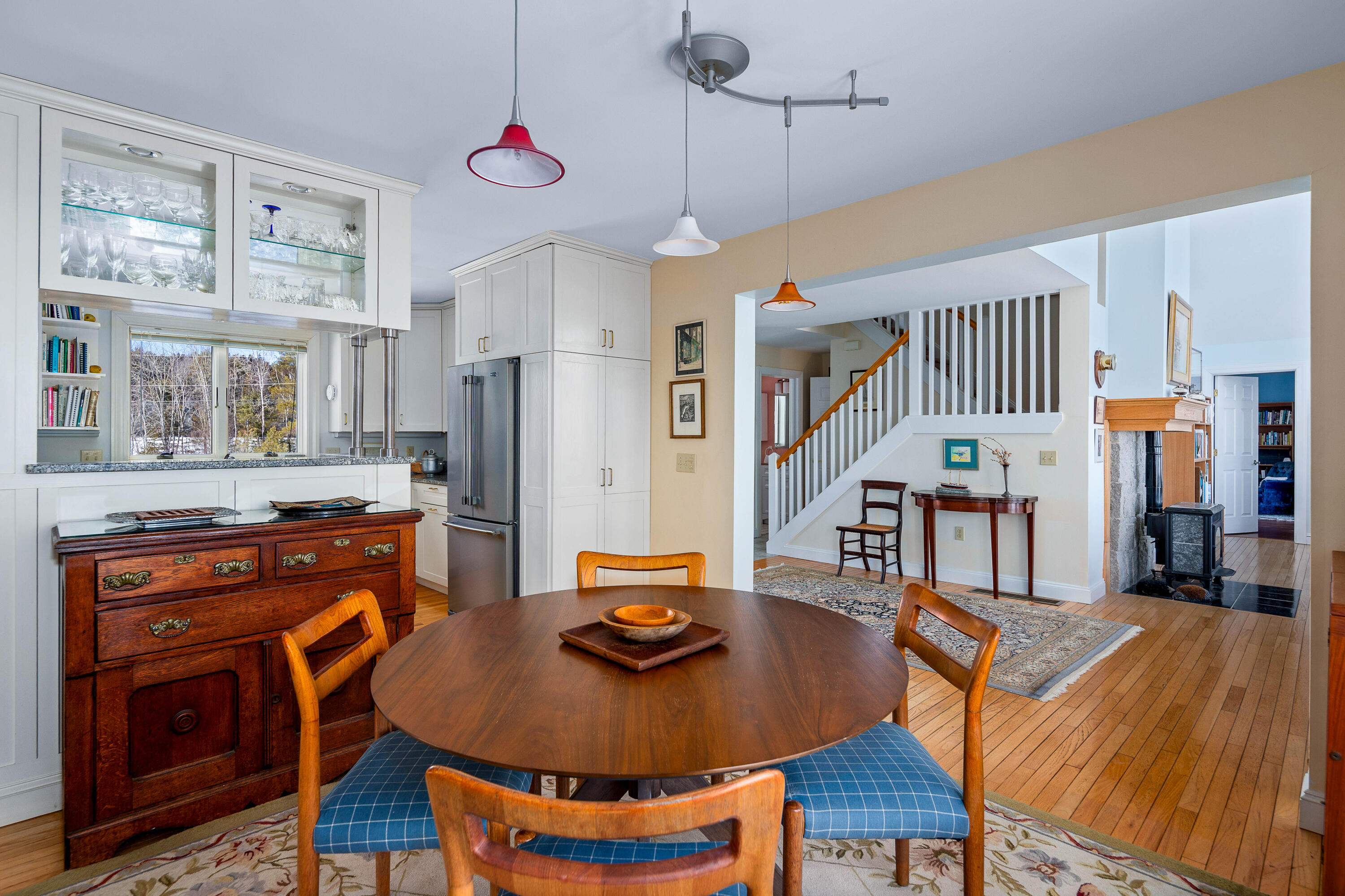 Open-plan dining room and kitchen with white cabinetry, a round wooden table, and a staircase beside a stone fireplace.