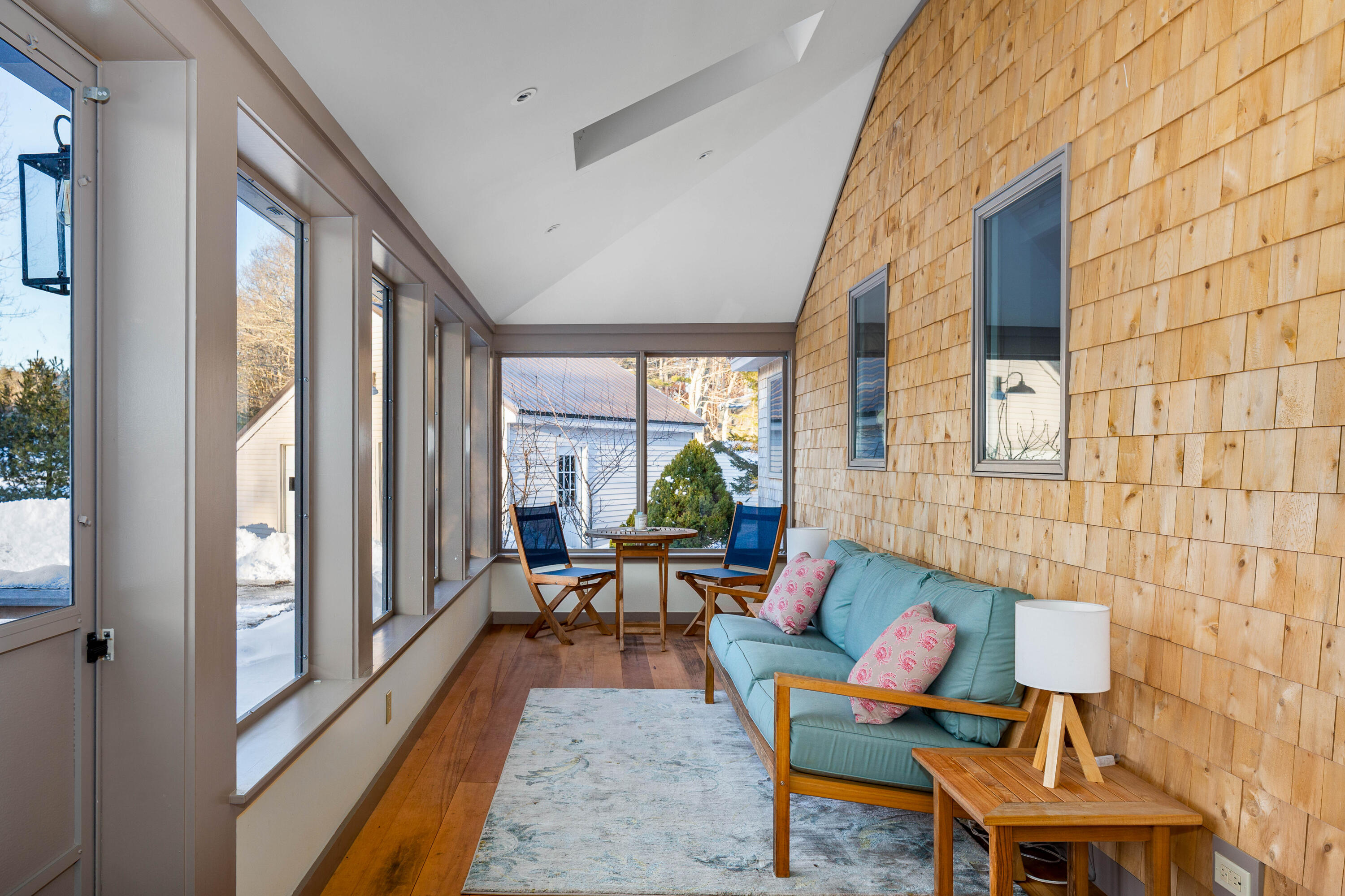 Furnished sunroom with a teal sofa, honey-colored shingle walls, and tall windows overlooking snow.