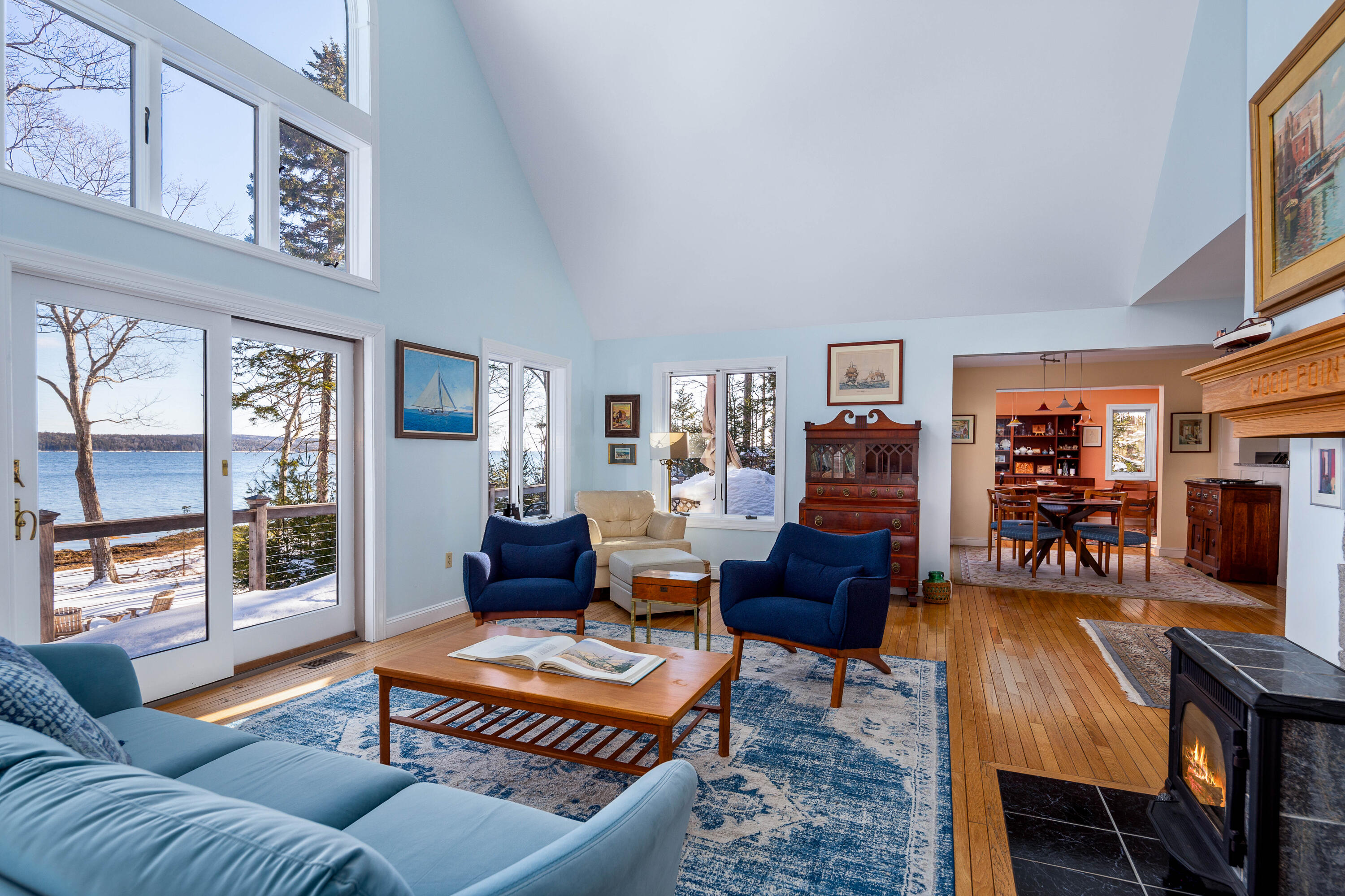 Living room with vaulted ceiling and blue seating, overlooking snowy water through large windows.