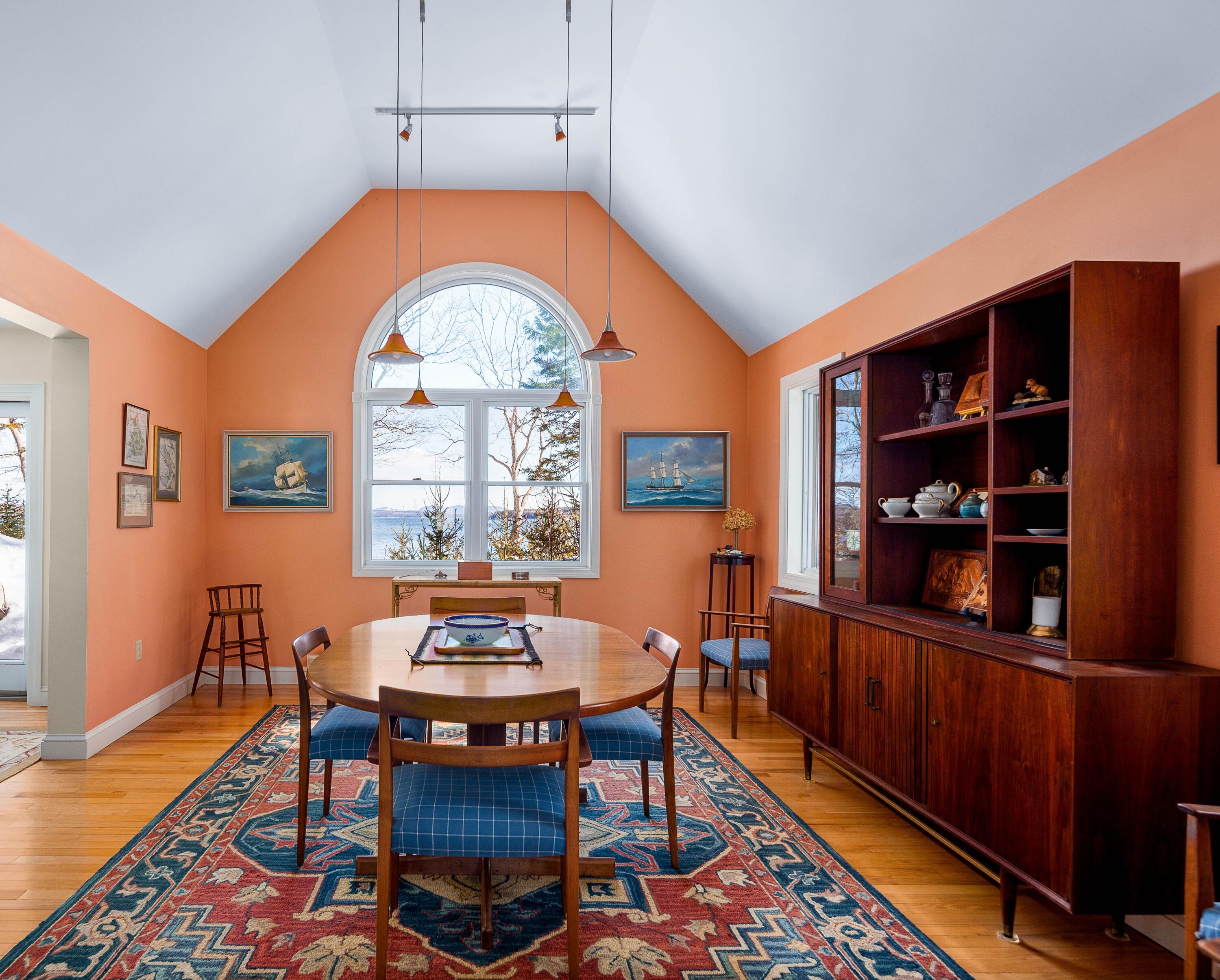 Dining room with a vaulted ceiling, arched water-view window, wooden table, and large display cabinet.