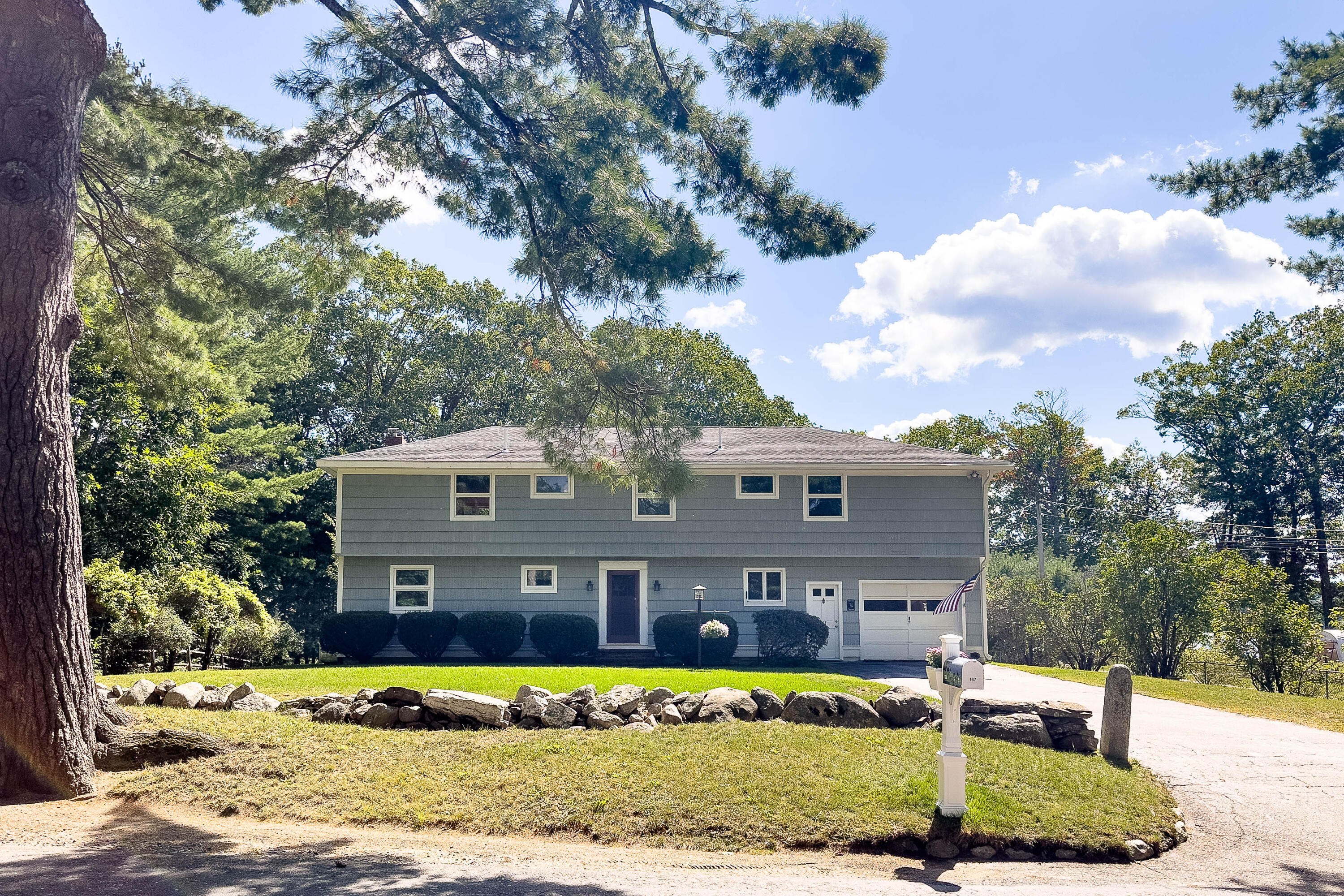 Gray two-story home with an attached garage, framed by mature trees and a stone border.