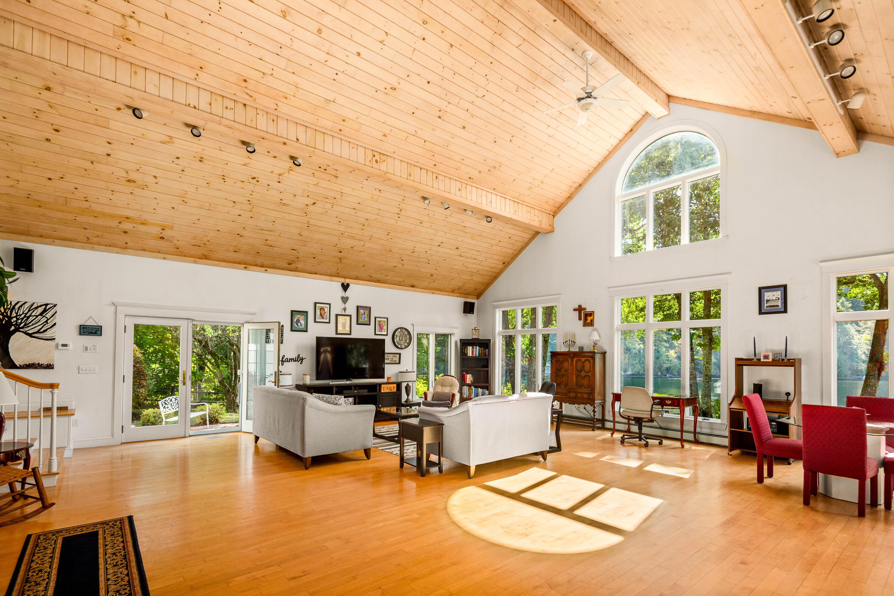 Bright living room with vaulted pine ceiling, tall arched window, and lake views through glass doors.