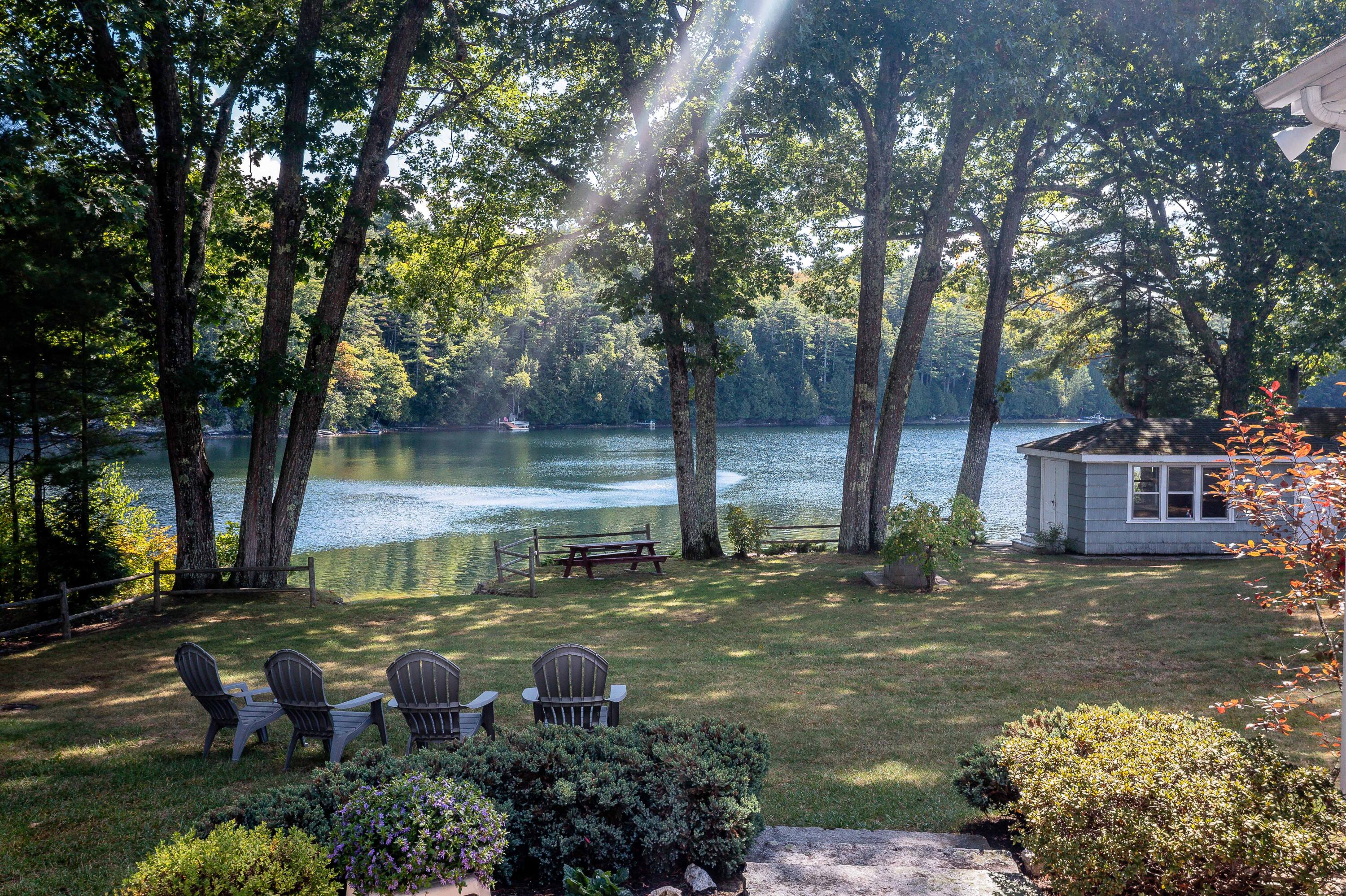 Shaded lawn with Adirondack chairs overlooking a wooded lake, beside a small lakeside building.