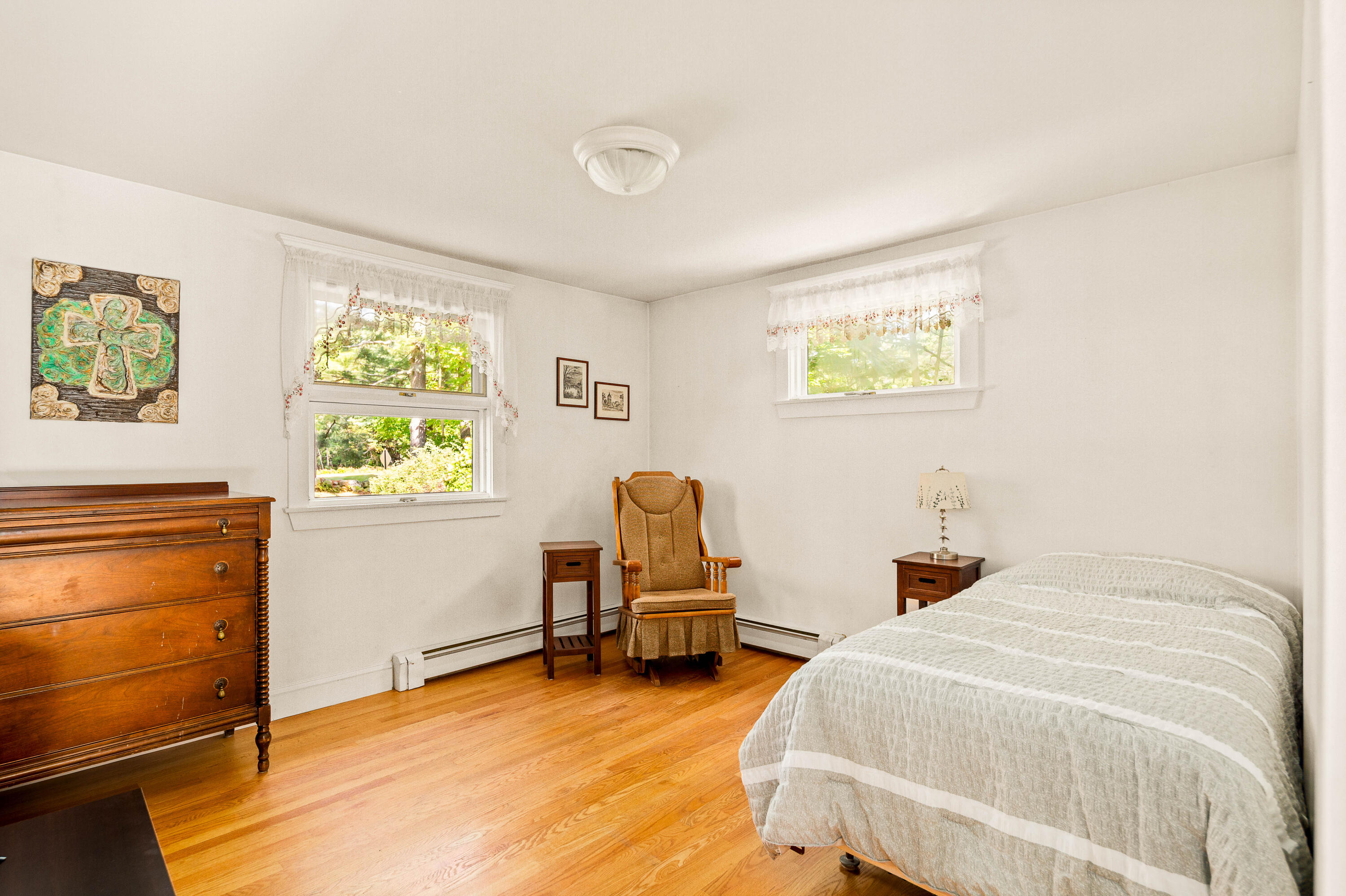 Bedroom with a white-covered bed, antique wooden dresser, and upholstered armchair beside sunny windows.