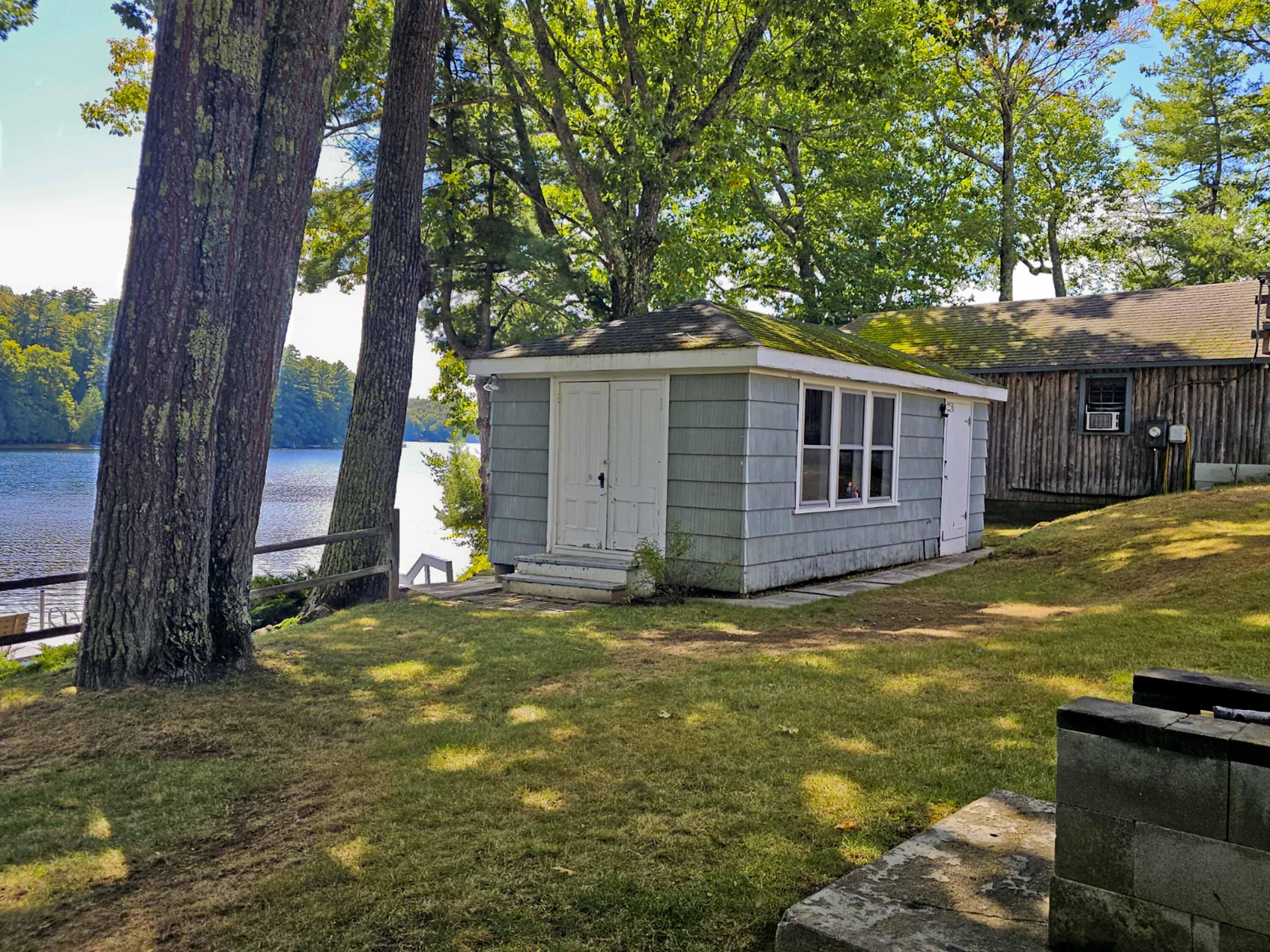 Small gray lakeside outbuilding beneath mature trees, with the pond visible behind it.