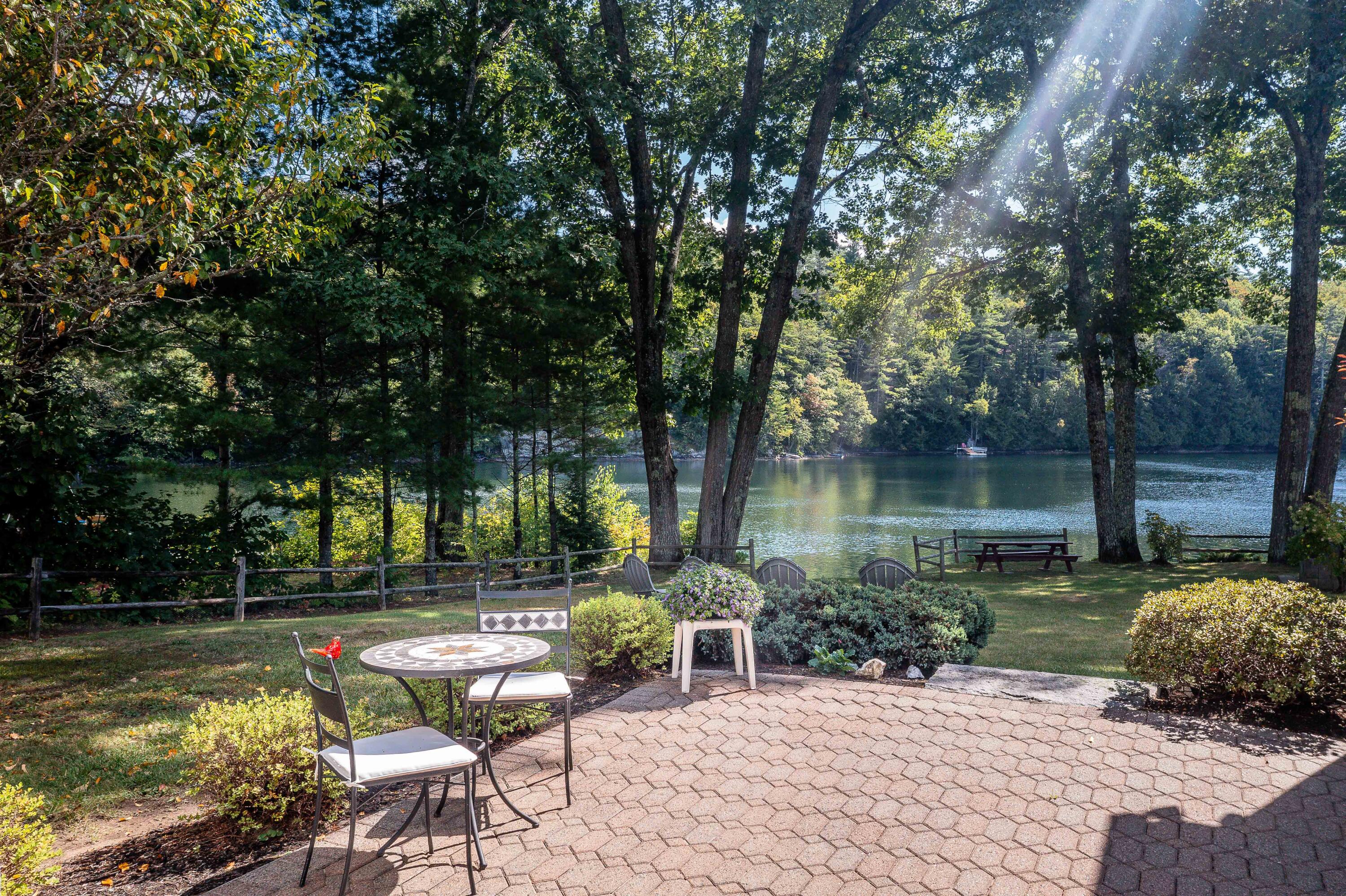 Paved lakeside patio with café table, Adirondack chairs, and a wooded pond view