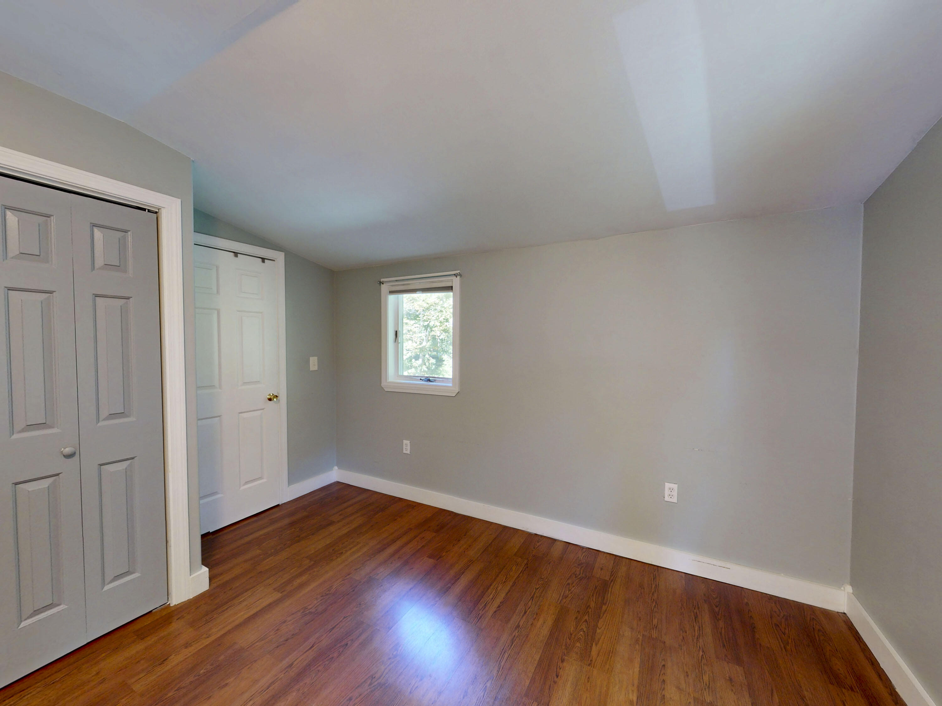 Empty room with reddish hardwood floors, gray walls, a small window, and white doors.