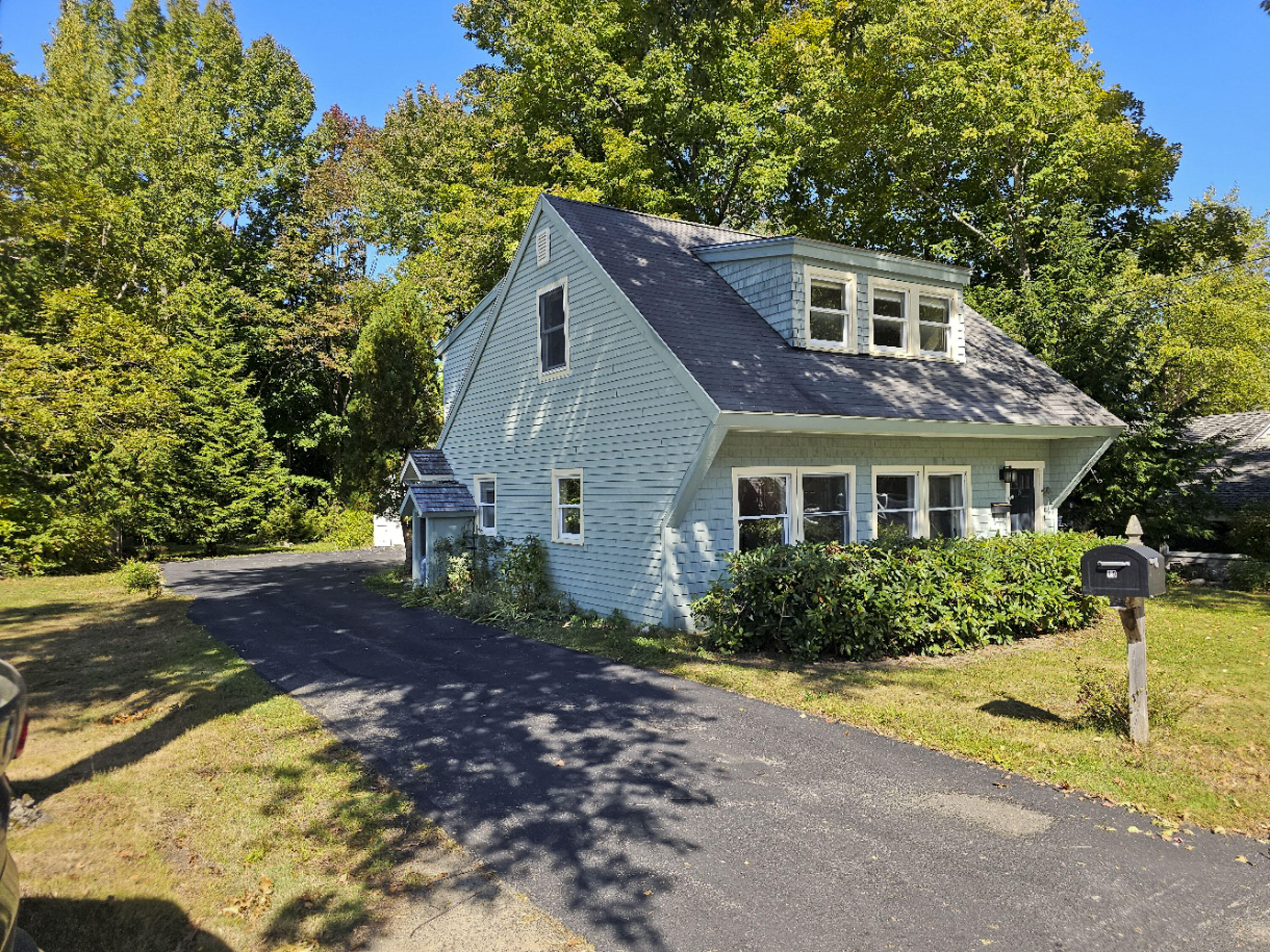 Light blue shingle-sided house with dormer windows, a curved driveway, and mature trees.