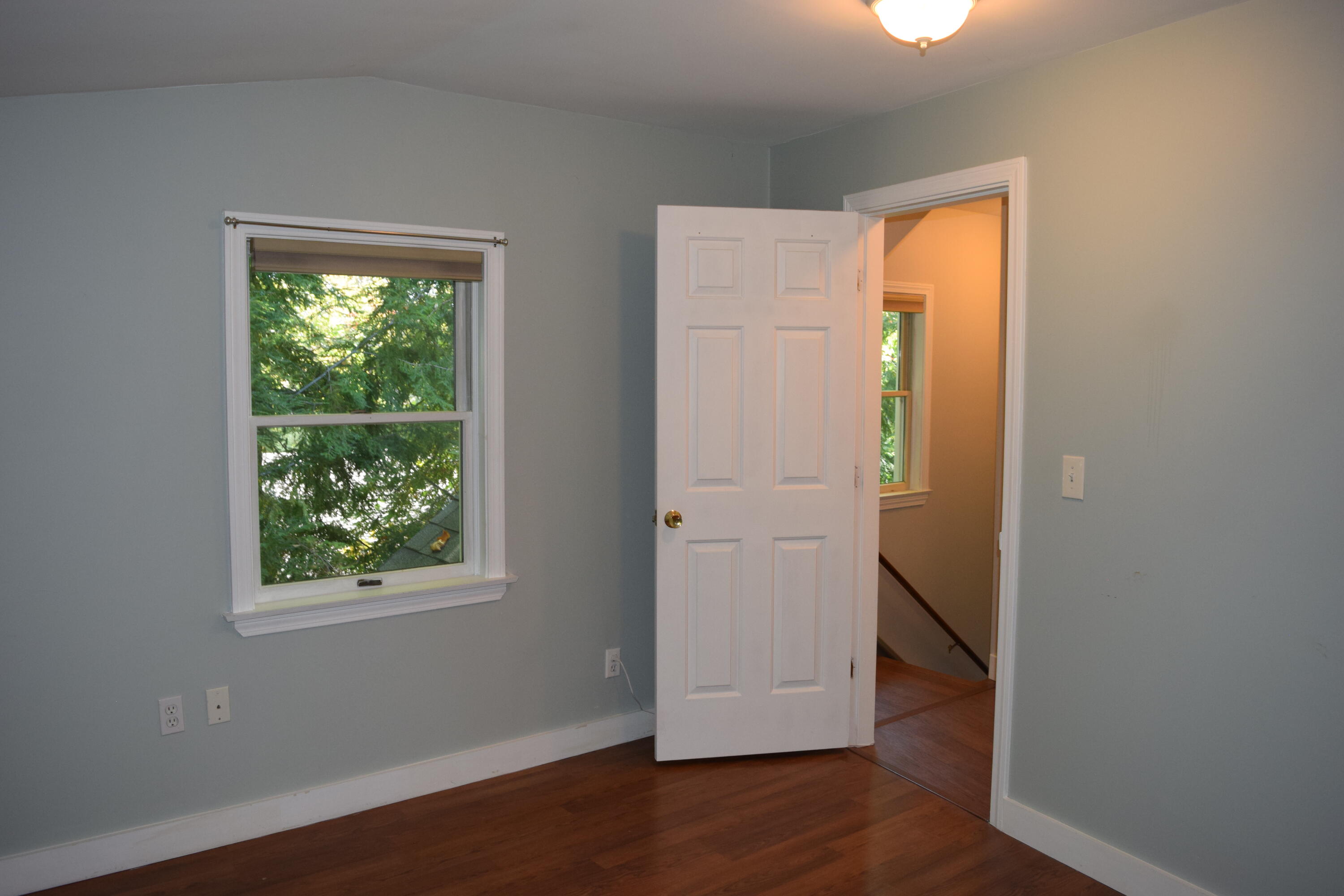 Empty gray room with hardwood floors, a leafy view through the window, and an open door to the hall.