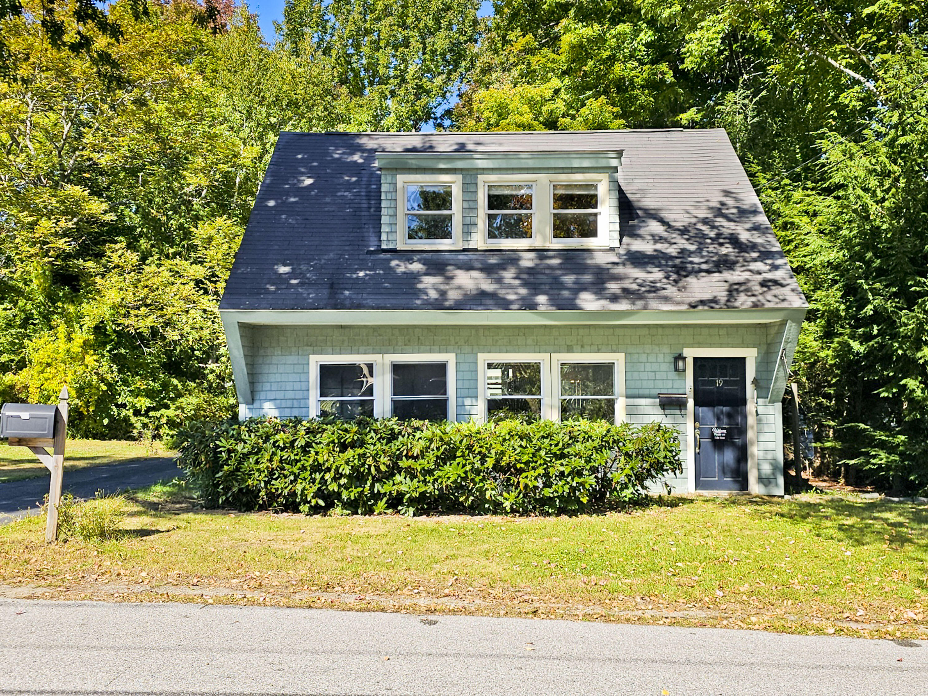 Pale blue-green shingle house with a steep roof, dormer windows, and front hedge among autumn trees.