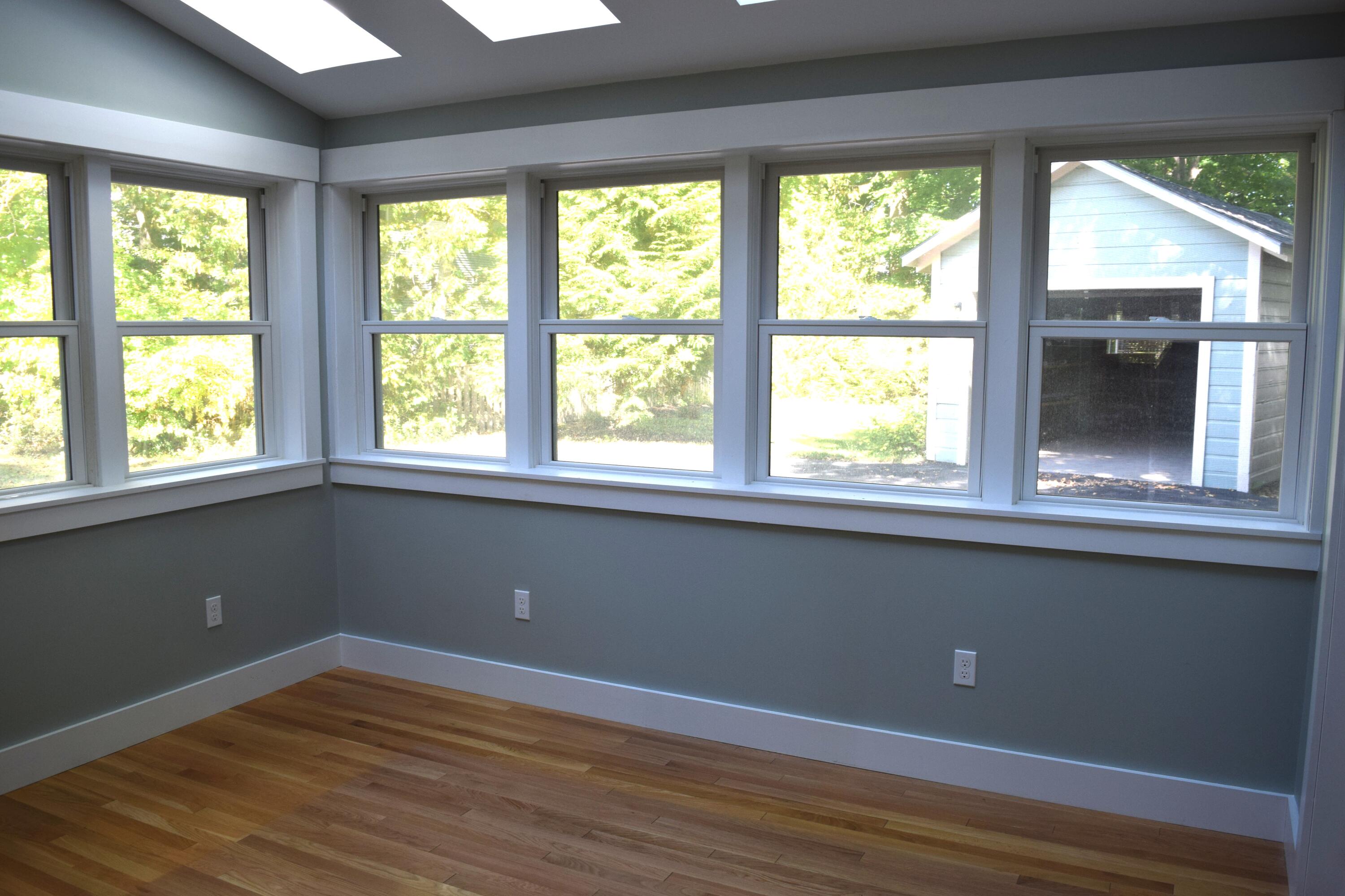 Empty sunroom with hardwood floors and large windows overlooking trees and a detached garage.