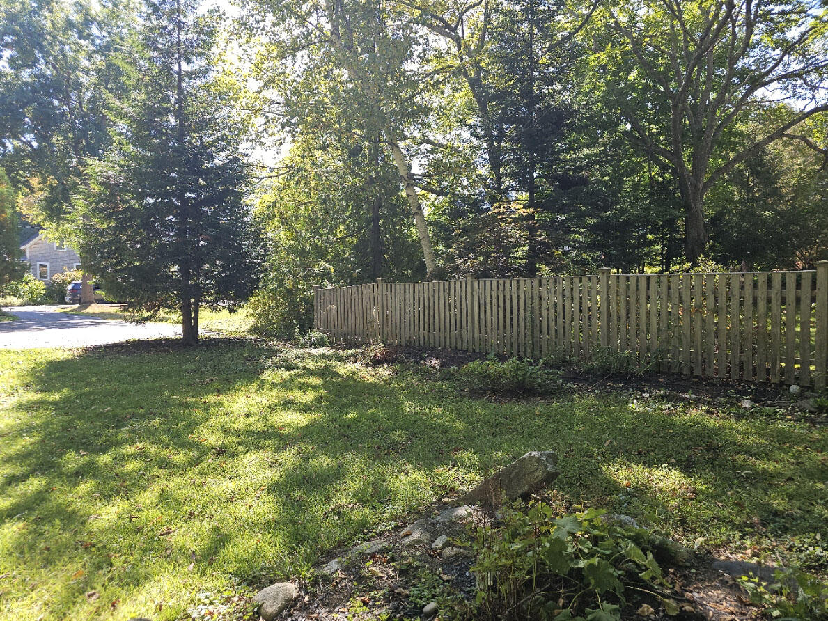 Tree-shaded yard with a wooden picket fence, green lawn, and rocks in the foreground.