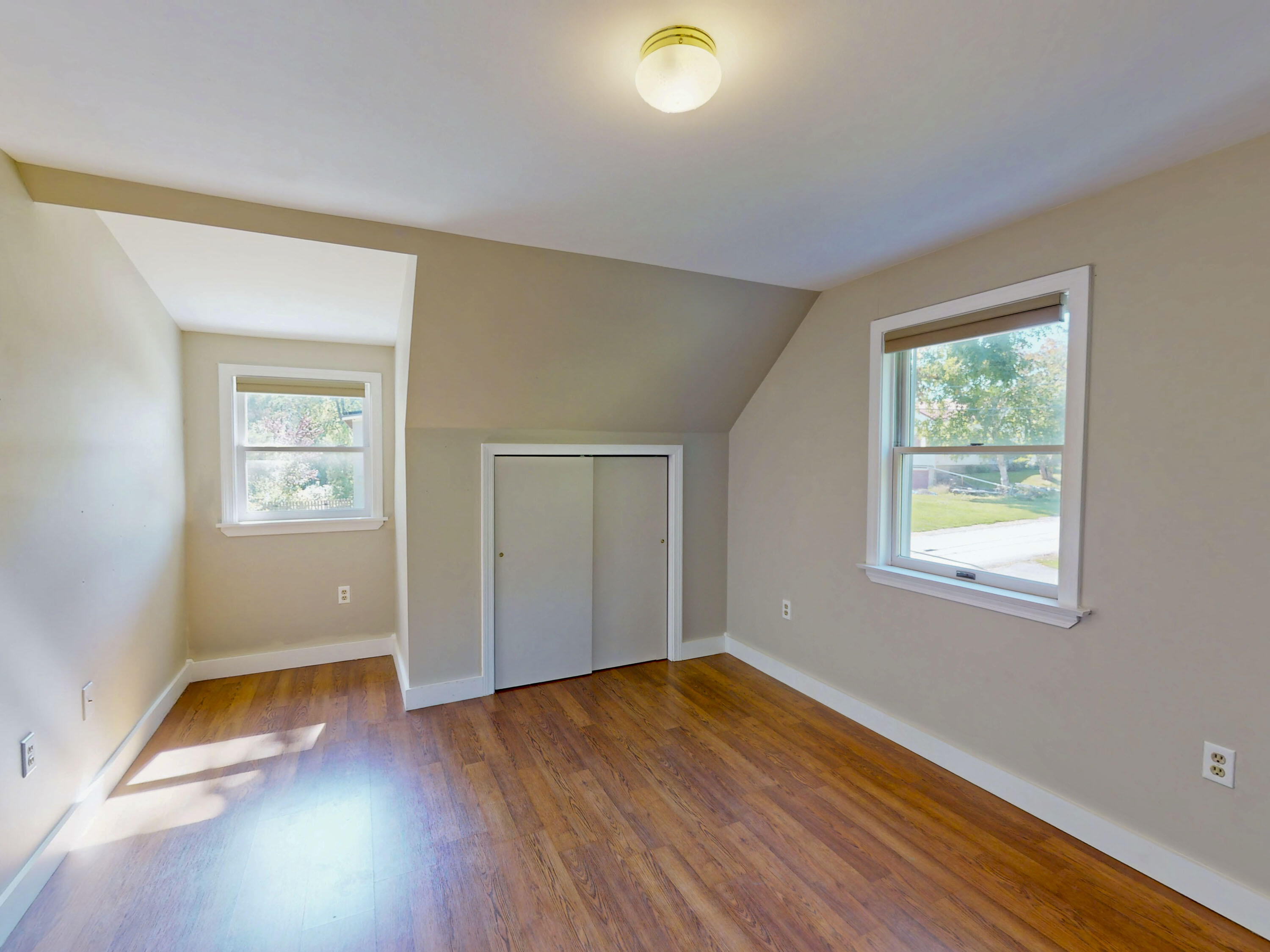 Empty bedroom with sloped ceilings, a sliding-door closet, two windows, and wood flooring.