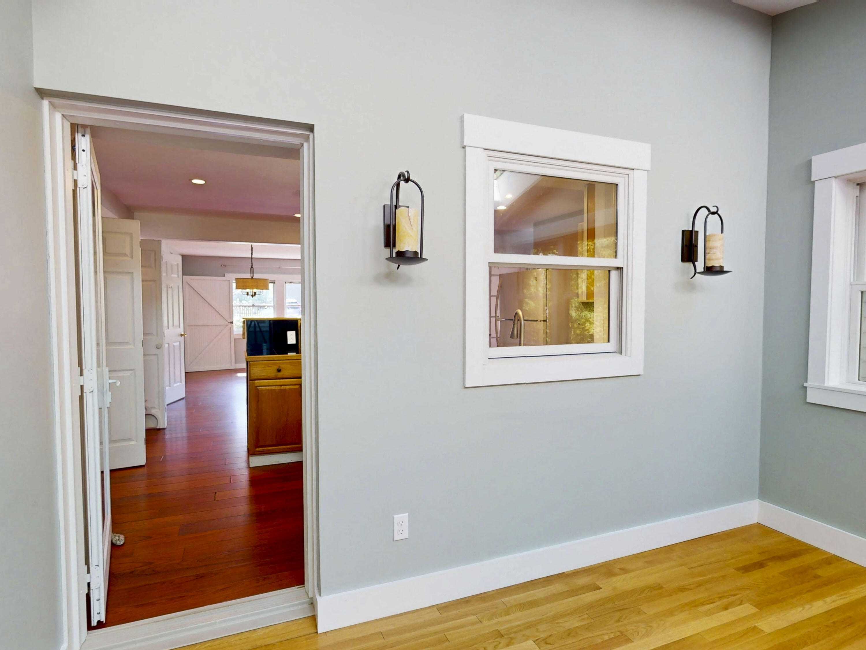 Light blue-gray room with hardwood floors, two wall sconces, and an open doorway to an adjoining room.