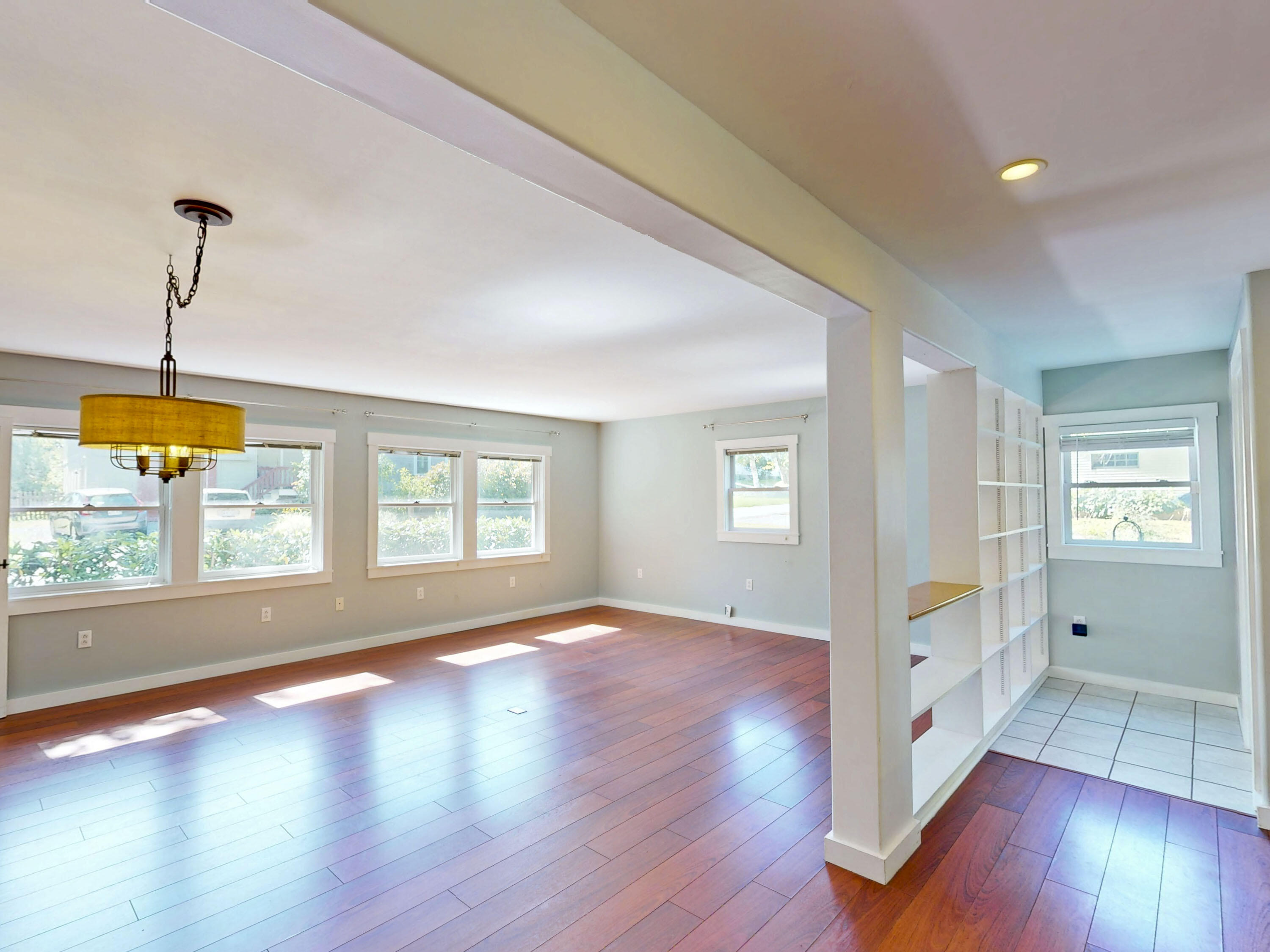 Vacant living area with reddish hardwood floors, broad windows, and built-in shelving beside a tiled nook.