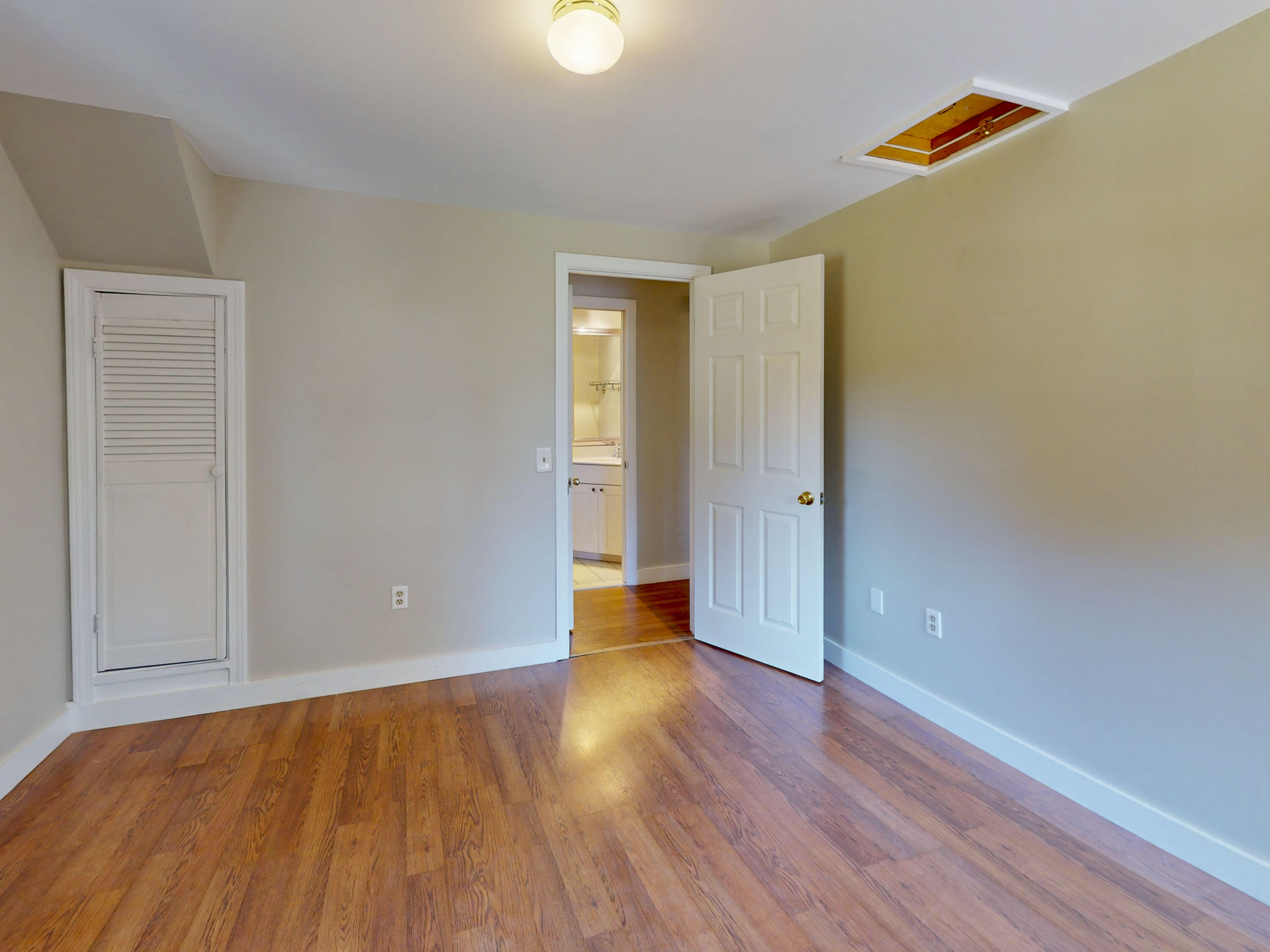 Empty bedroom with hardwood floors, a louvered closet door, and an open bathroom doorway.