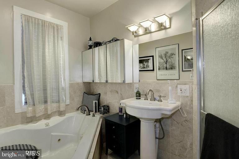 Bathroom with a jetted corner tub, pedestal sink, marble-look tile, and mirrored medicine cabinets.