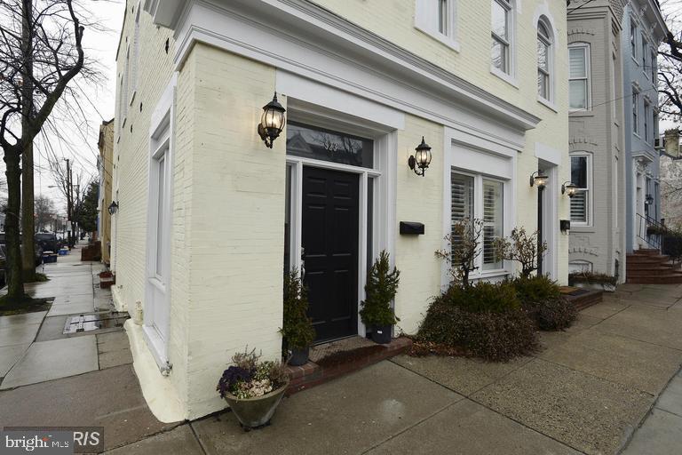 Pale yellow brick townhouse with a black front door and lanterns along a wet sidewalk.