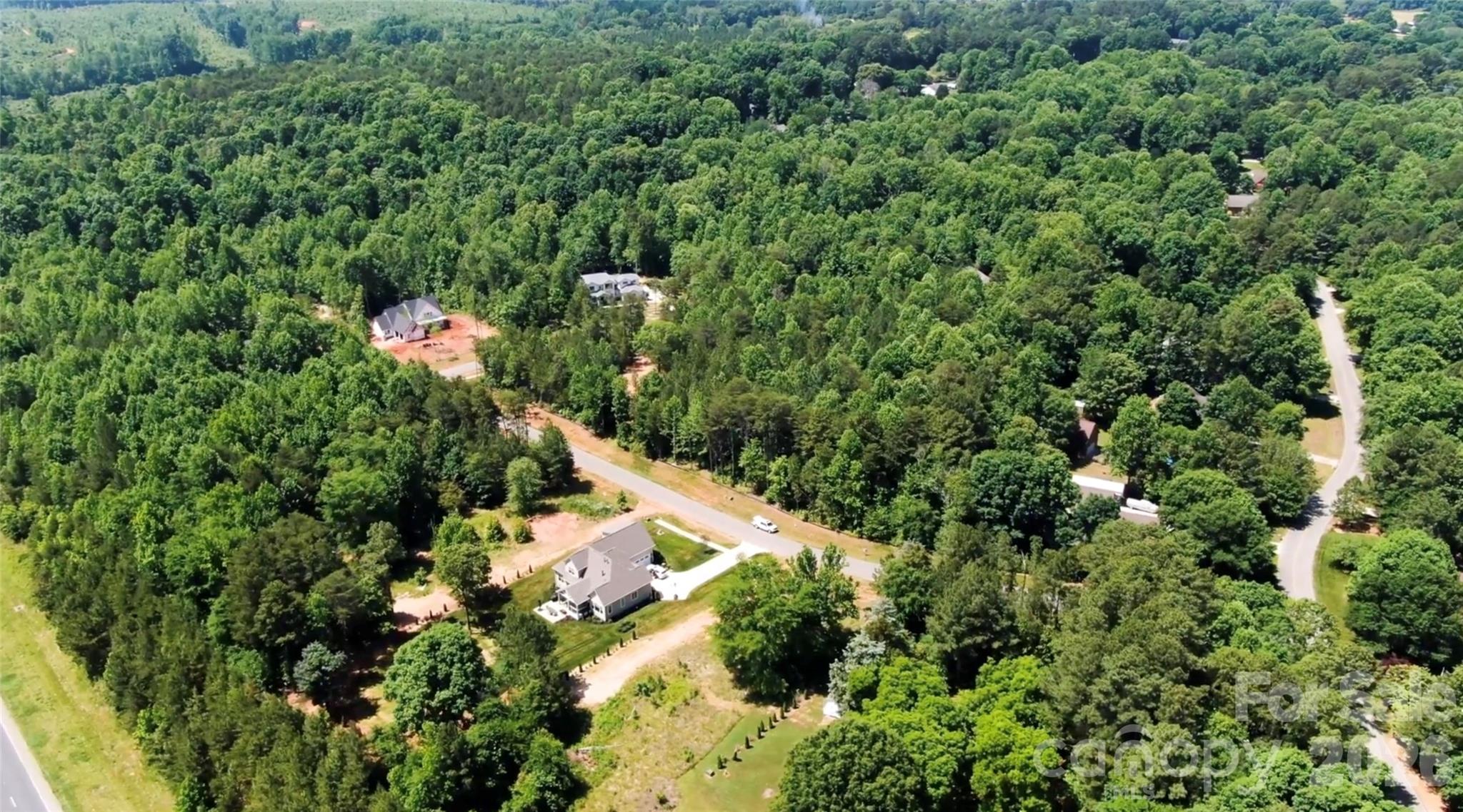 Aerial view of a gray two-story home beside a road amid dense woodland and scattered neighboring houses.