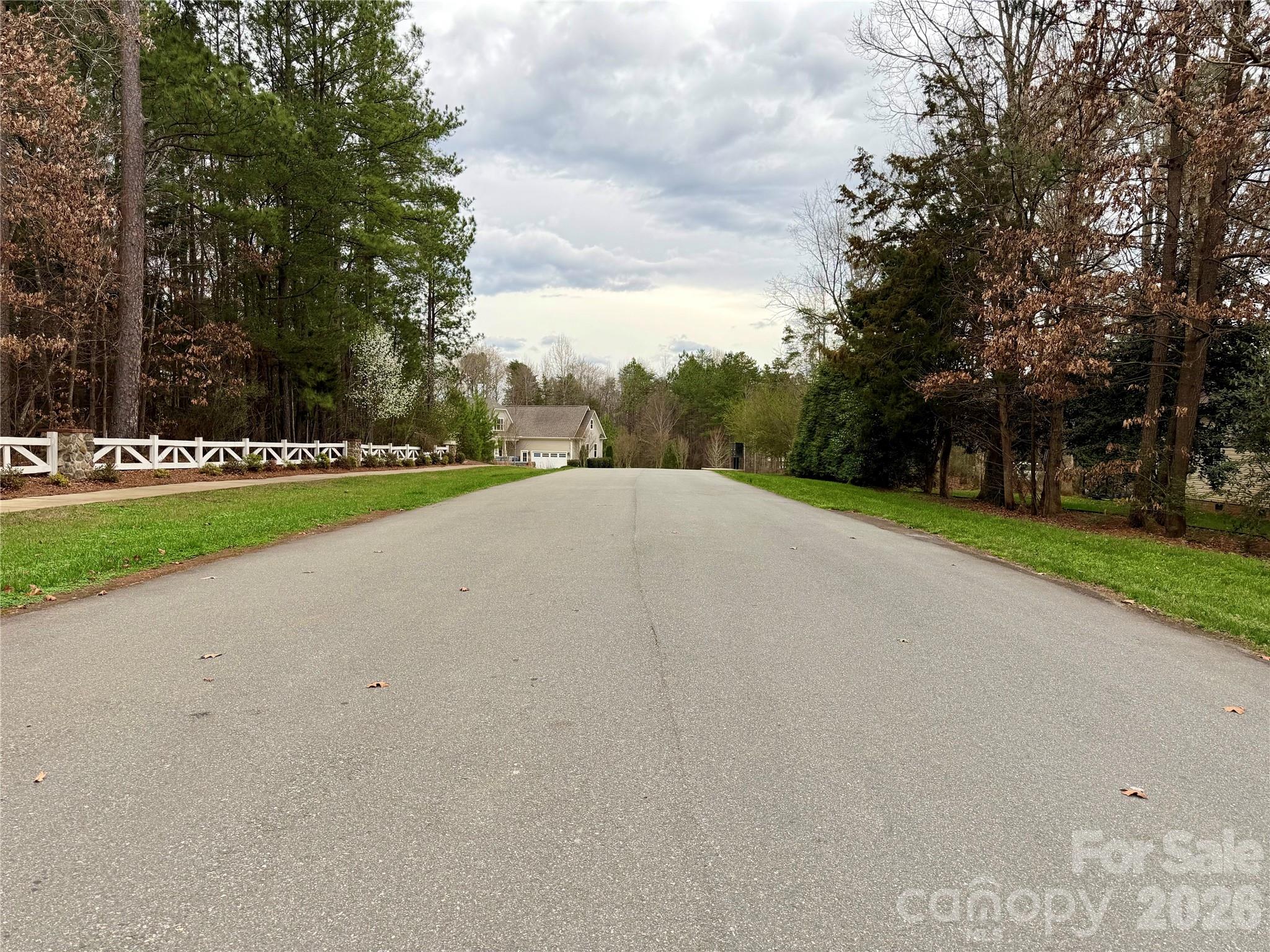 Paved residential road leading to a house, bordered by a white fence and trees.